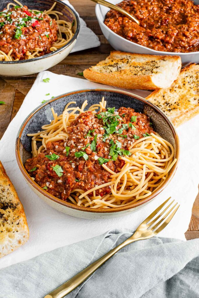 Side view of red lentil ragu poured overtop a bowl of spaghetti noodles. The ragu is sprinkled with parsley and garlic bread rests beside the pasta bowl.