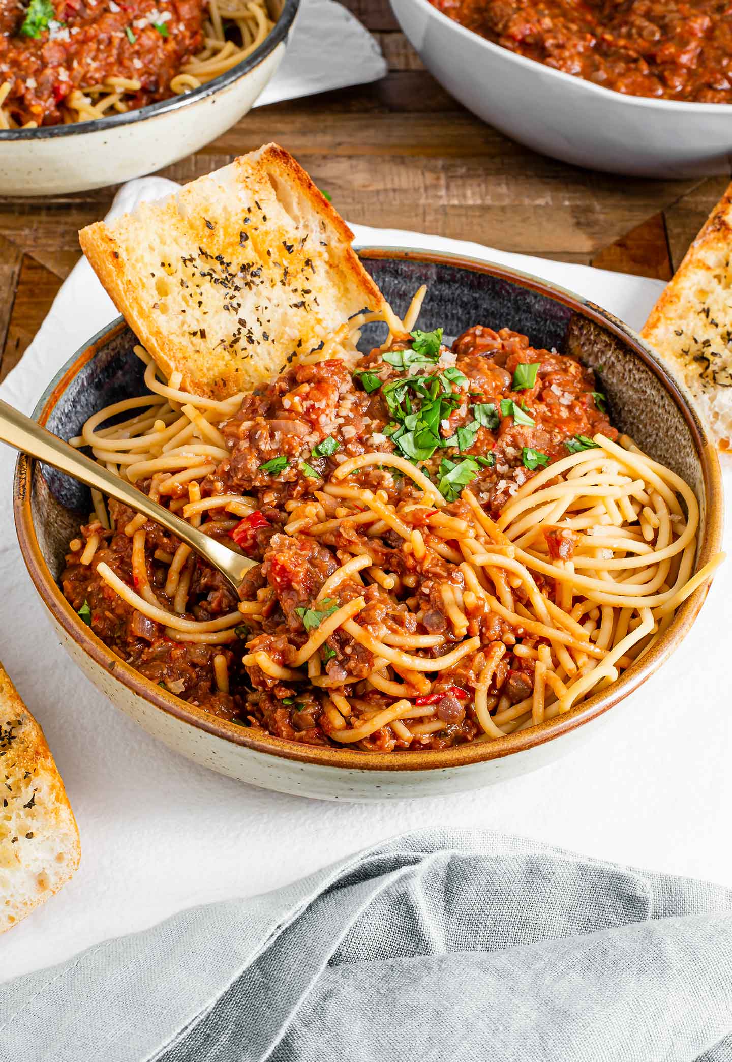 Side view of a bowl of lentil ragu served over spaghetti noodles. A slice of toasted garlic bread rests against the edge of the bowl.