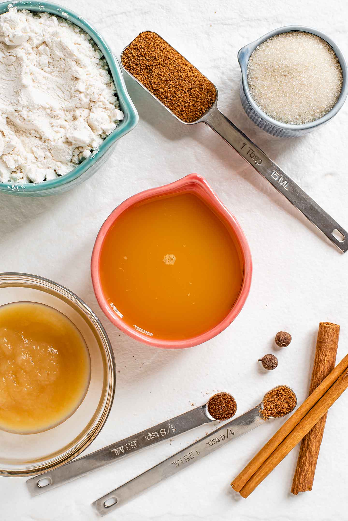 Top down view of apple cider, applesauce, white flour, coconut sugar, cane sugar, cinnamon sticks, allspice berries, ground cloves, and ground nutmeg.