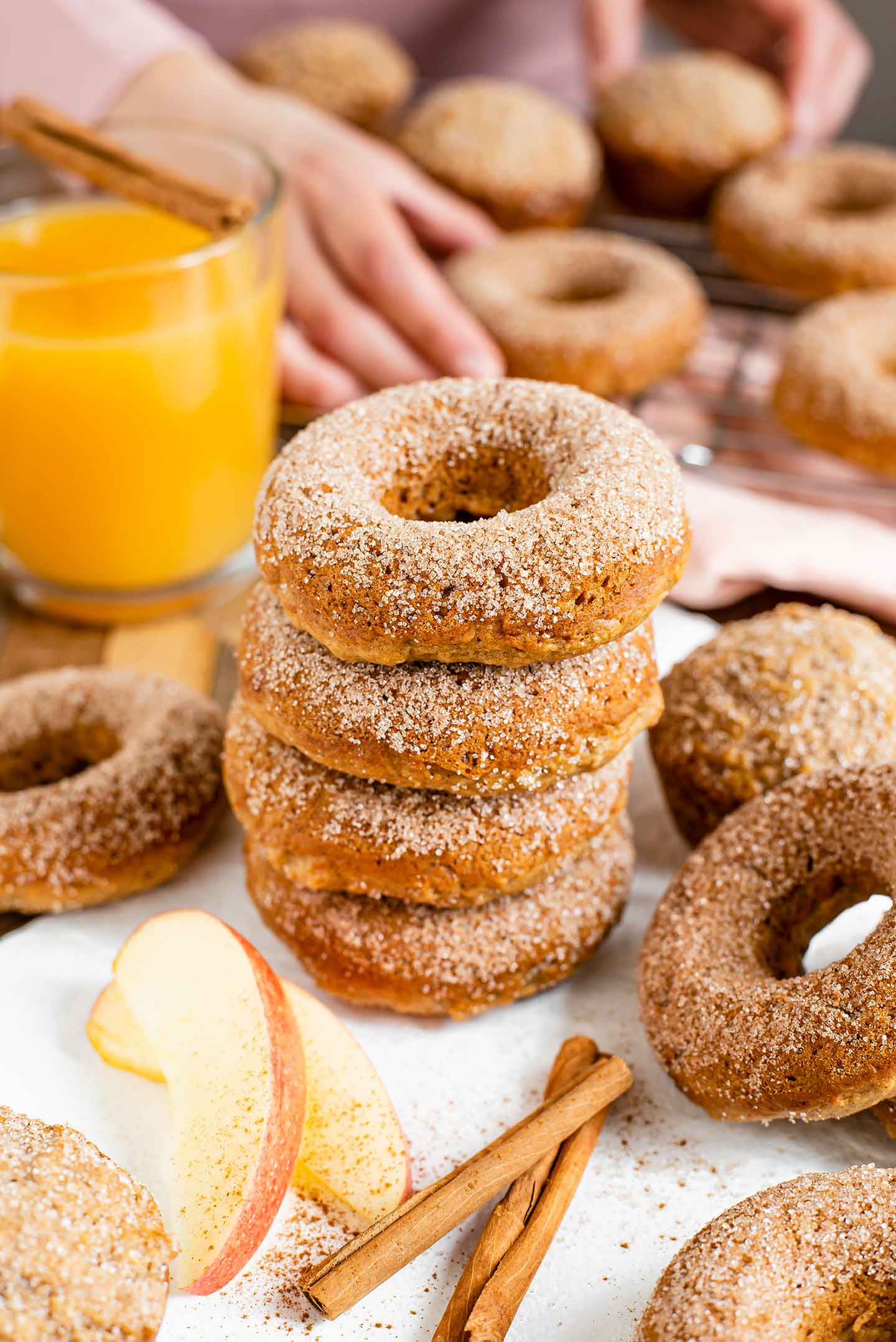 Side view of apple cider doughnuts stacked on a white tray. The top of the doughnuts are sprinkled with cinnamon sugar, the remaining doughnuts are scattered around, and apple cider muffins can also be seen.