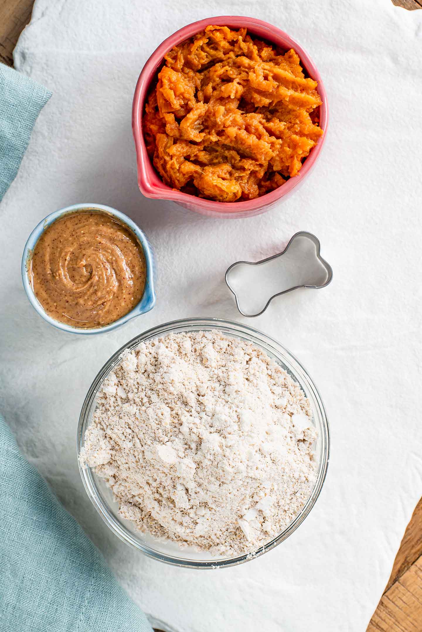 Top down view of ingredients on a white tray. Mashed sweet potato fills a small bowl next to small bowls of peanut butter and oats ground into a fine flour.