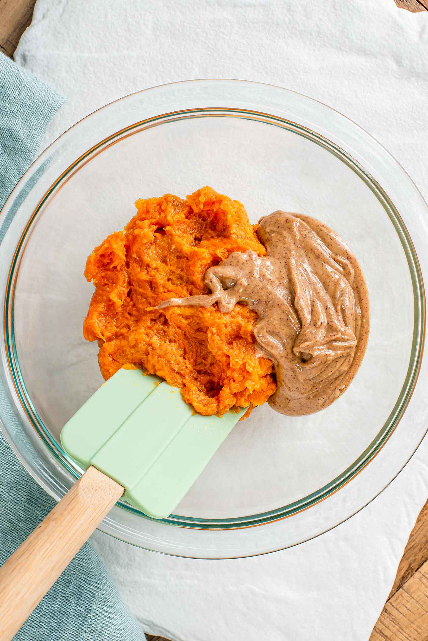 Top down view of mashed sweet potato and runny peanut butter in a glass bowl with a rubber spatula.