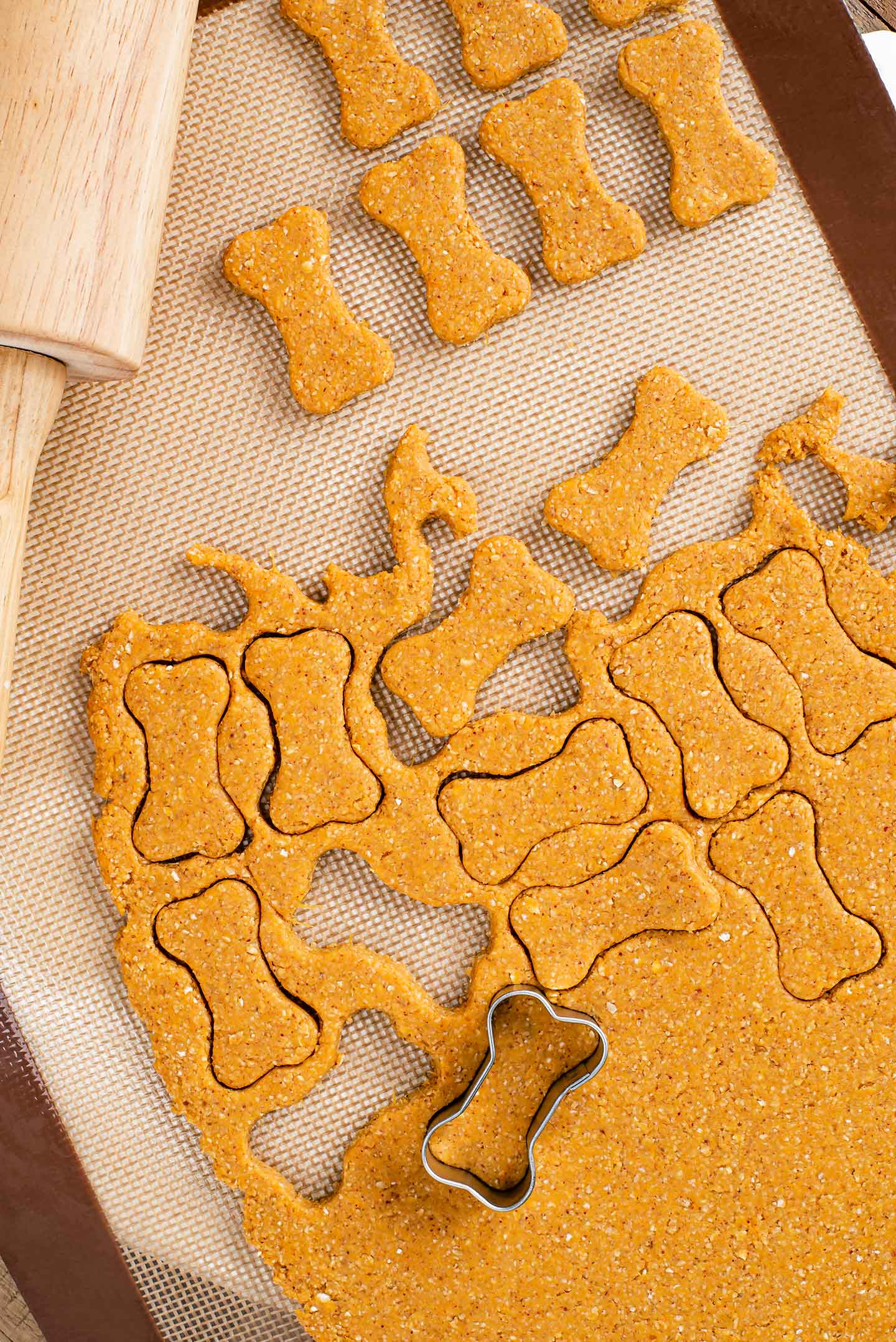 Top down view of rolled dog treat dough on a silicone mat with a rolling pin. A small dog bone cookie cutter has cut biscuit shapes into the dough.