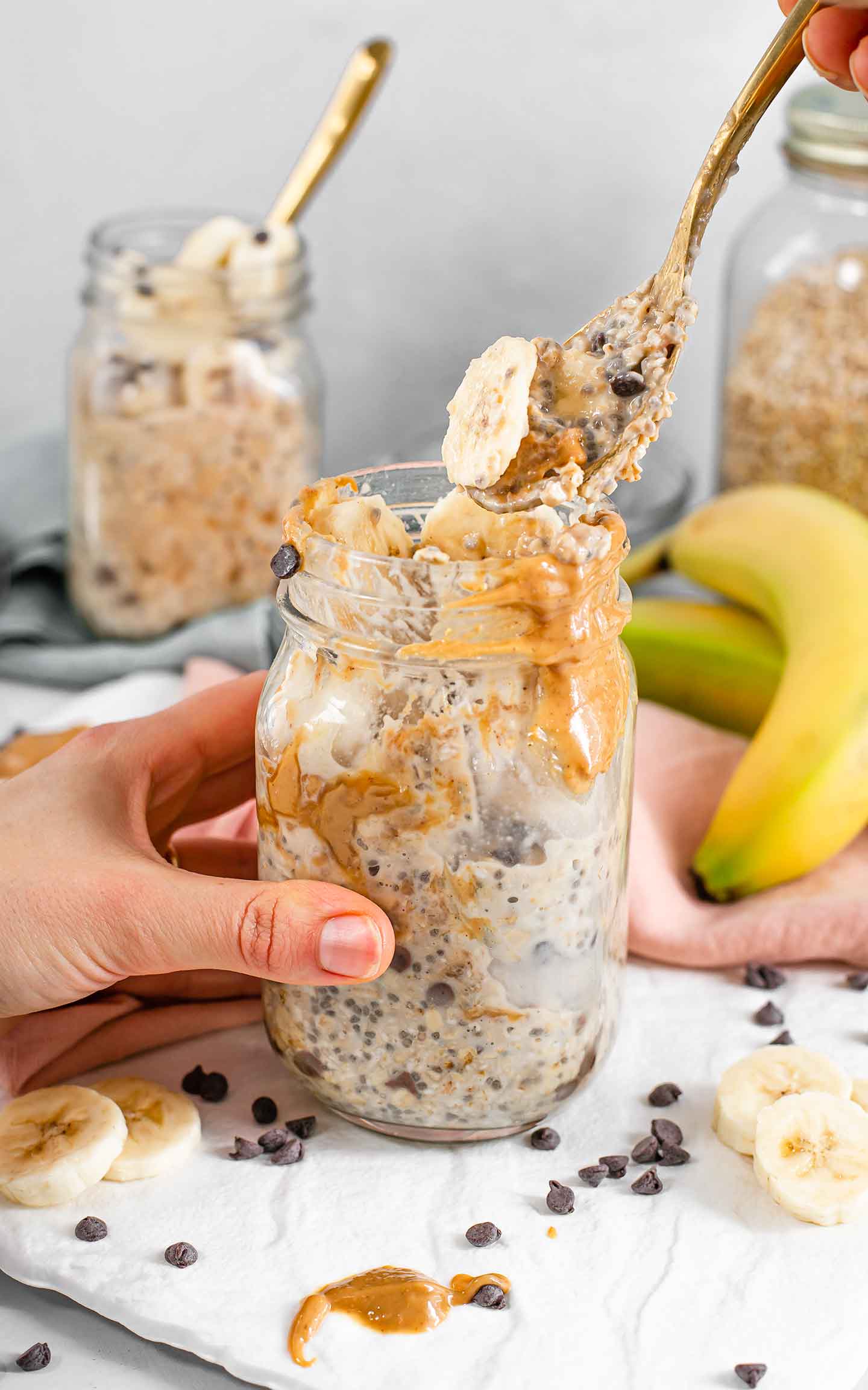 Side view of a hand holding a mason jar and digging into PB Banana Chip Oats with a spoon. Chocolate chips and banana slices are scattered around, another mason jar of PB Banana Chip Oats is in the background and a jar of oats is in the background.