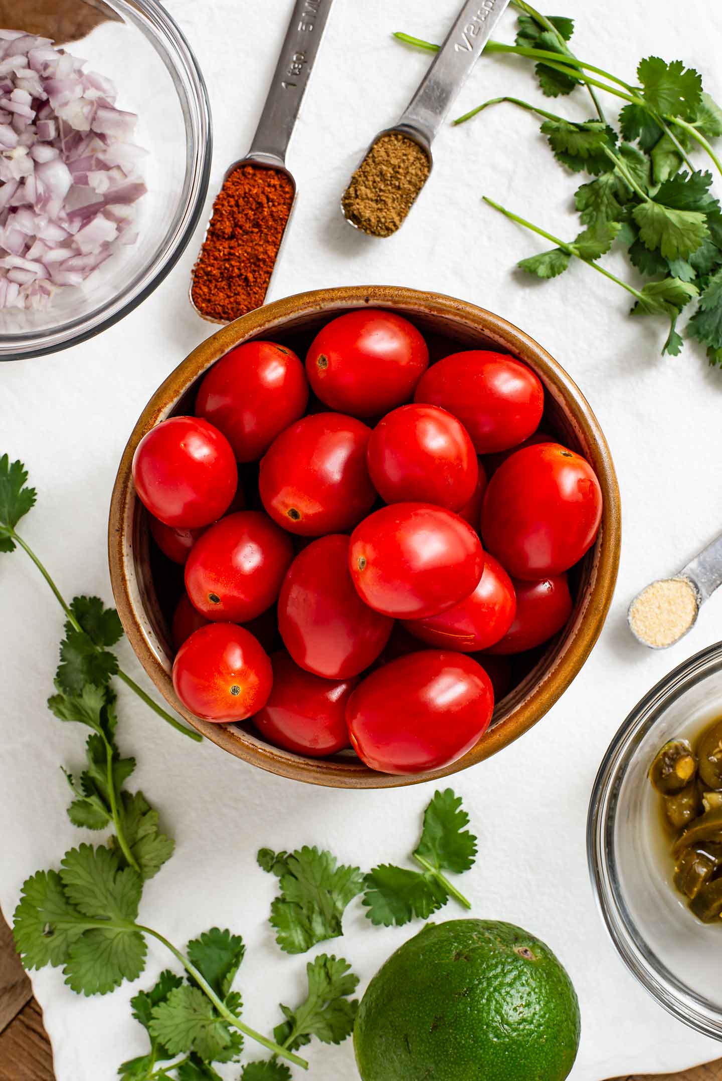 Top down view of grape tomatoes in a bowl with chopped red onion, fresh cilantro, a lime, sliced jalapeños, and seasonings all atop a white tray.