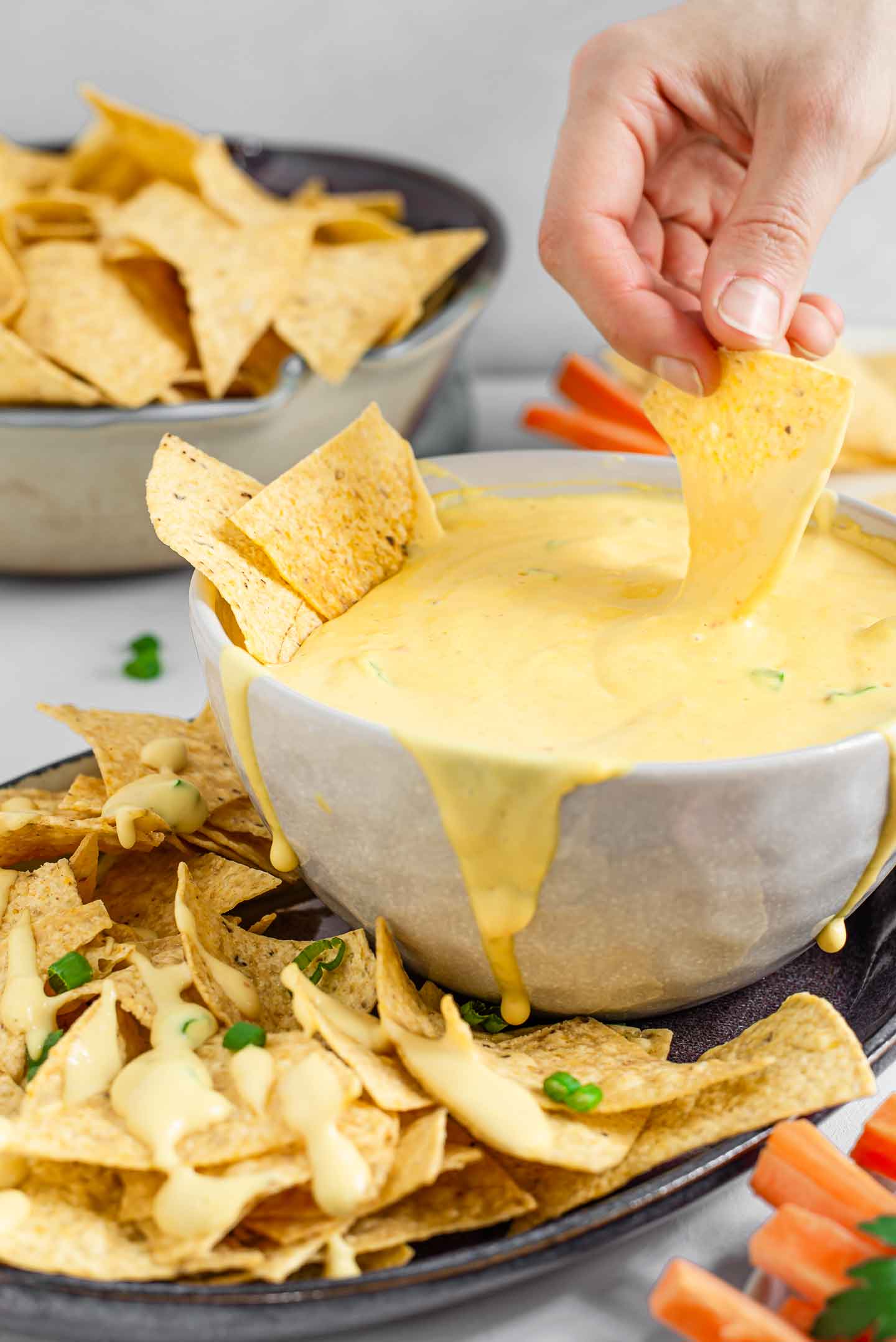 Side view of a hand dipping a tortilla chip into a large bowl of vegan queso. Queso drips down the side of the bowl and tortilla chips and carrot sticks surround it.