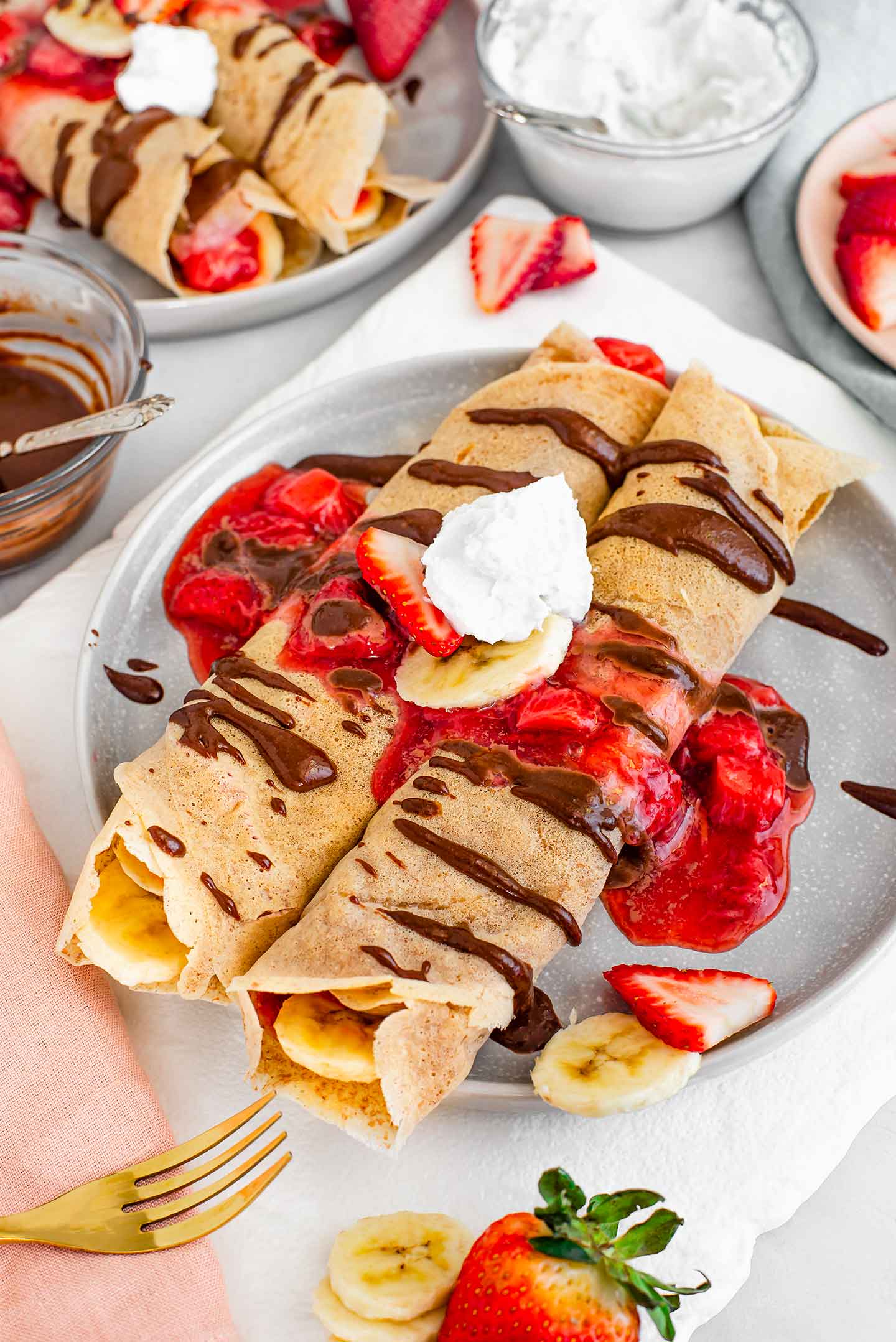 Side view of two crepes on a plate topped with strawberry sauce, chocolate drizzle and whipped cream. Another plate of crepes and more whipped cream are in the background.