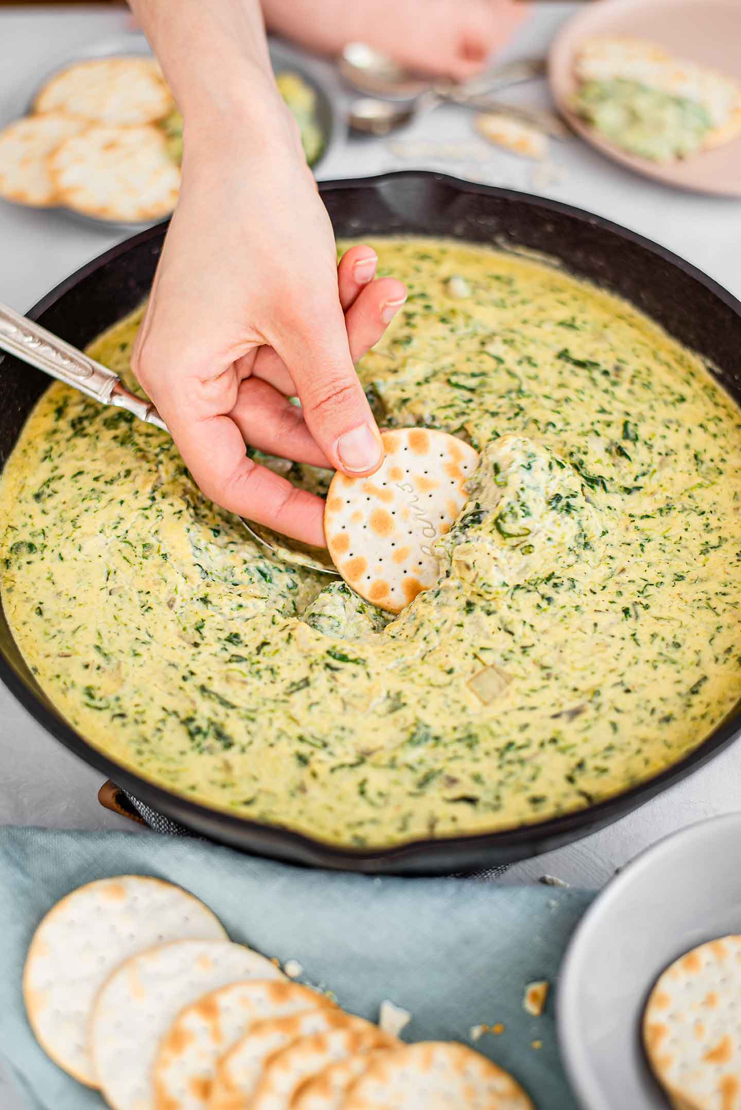 Side view of a hand dipping a cracker into the centre of a cast iron skillet filled with creamy spinach artichoke white bean dip.