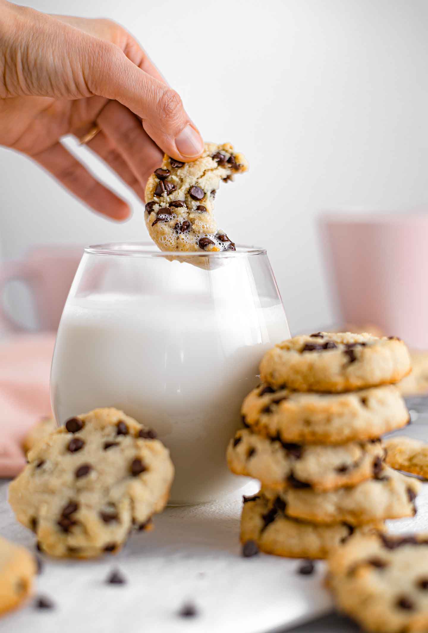 Side view of a chewy chocolate chip cookie with a bite taken out being dipped into a glass of milk. Other cookies are stacked against the glass and scattered around.