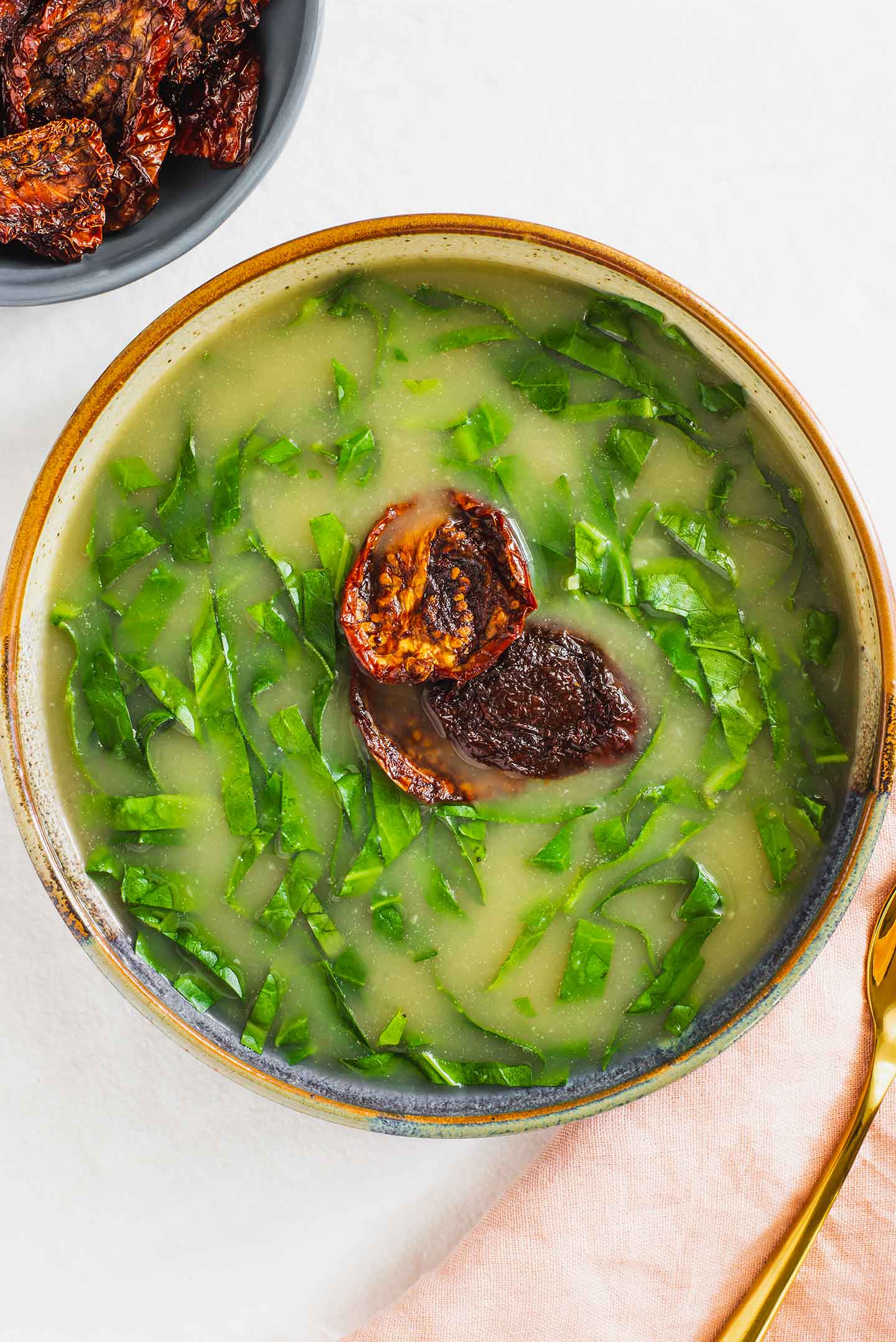 Portuguese caldo verde, collard soup, fills a wide shallow bowl. Shredded collard float in a thickened broth while three pieces of smoky sun-dried tomato decorate the center.