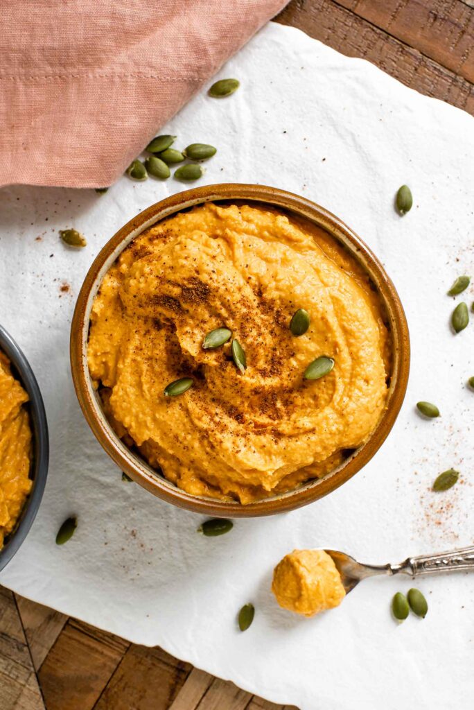 Top down view of an orange coloured pumpkin hummus filling a small bowl. The creamy hummus is garnished with pumpkin seeds and a sprinkling of nutmeg.
