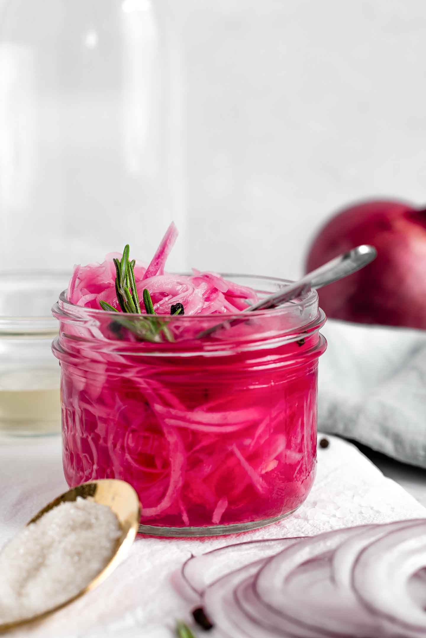 Side view of quick pickled red onions in a glass jar with vinegar, sugar, salt, and a whole red onion on a white tray.