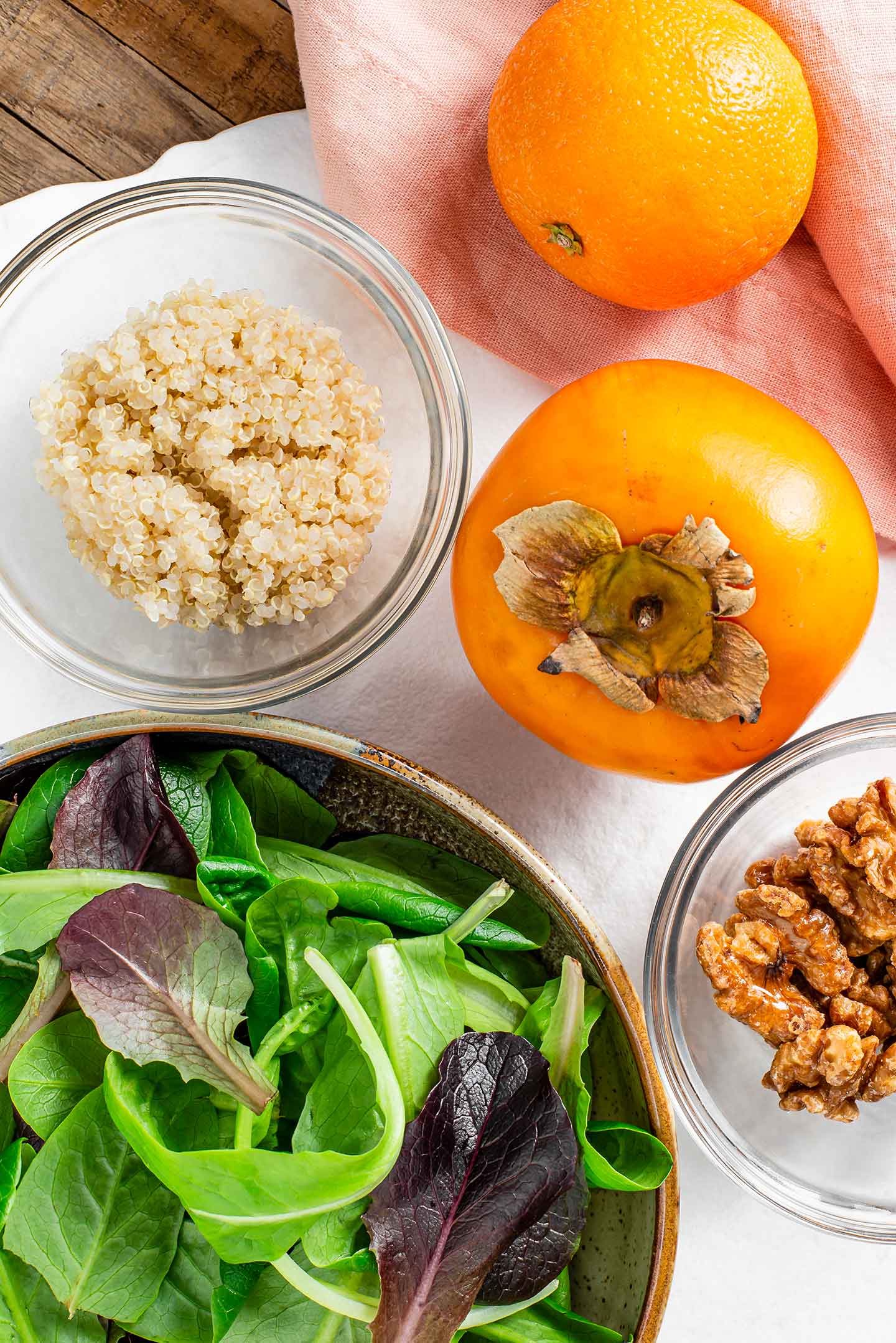 Top down view of a persimmon, an orange, small bowls of golden quinoa and candied walnuts, and a larger bowl of mixed greens.