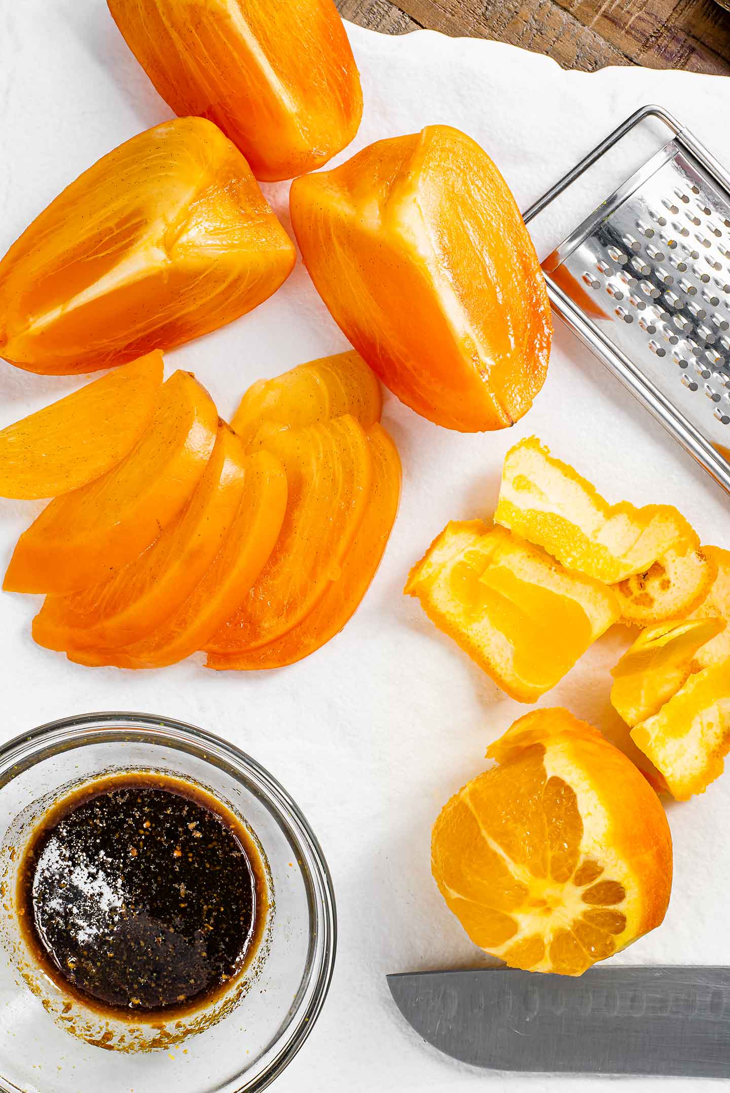 Top down view of sliced orange and persimmon on a white tray with a small dish of dark coloured dressing speckled with fresh orange zest.