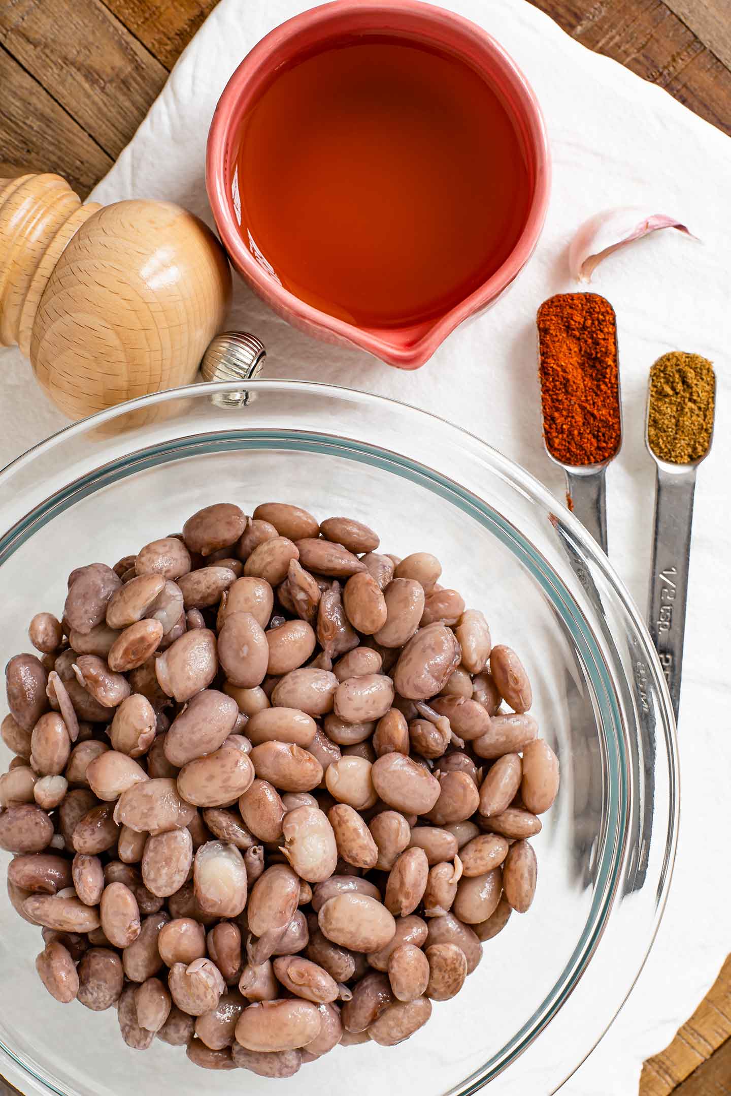 Top down view of pinto beans in a glass bowl. Arranged around the beans are seasonings, a clove of garlic, and a small bowl of broth.
