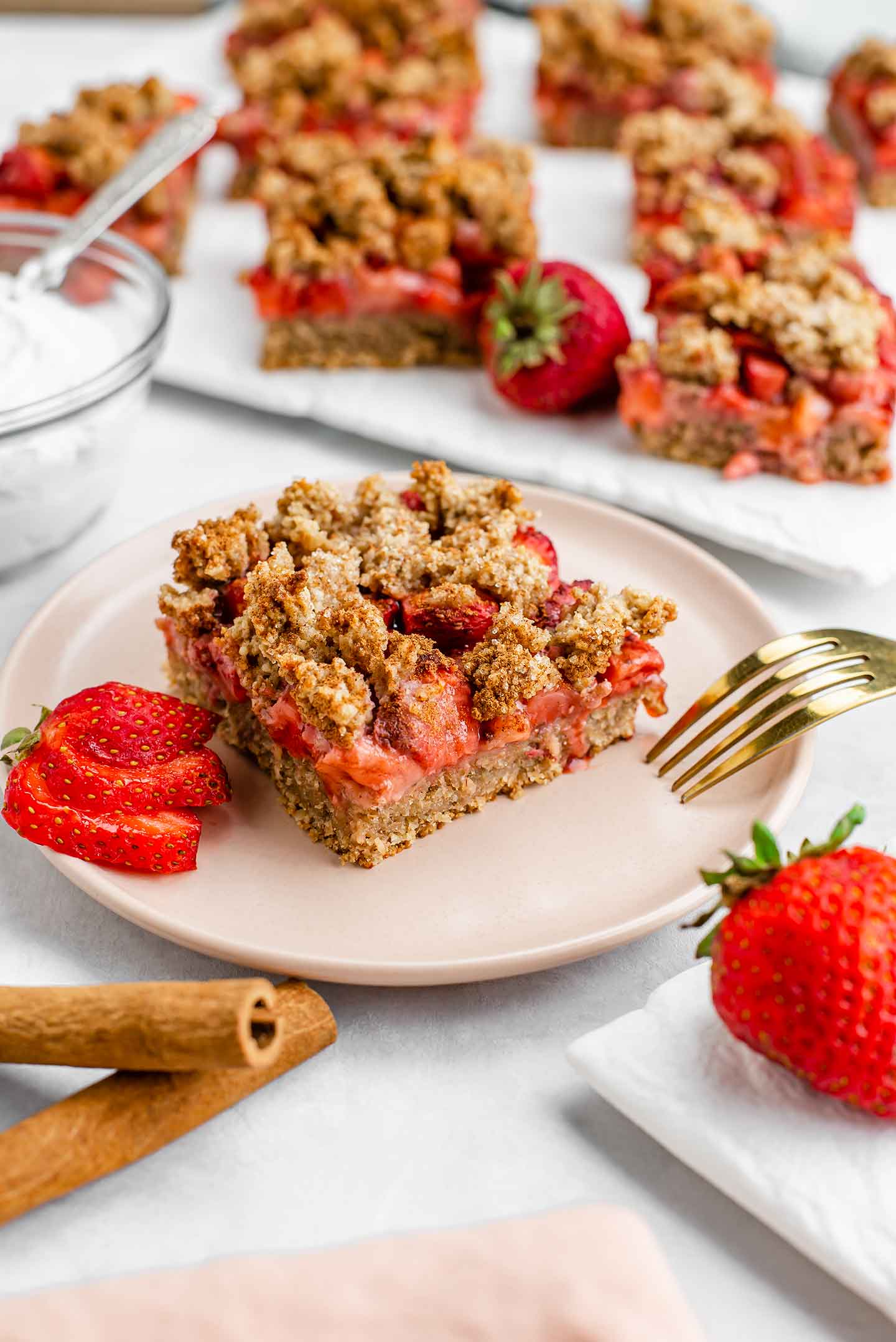 Side view of a strawberry crisp bar on a small plate. A tray of other bars is in the background. The crust is firm, the filling is gooey, and the topping is golden.