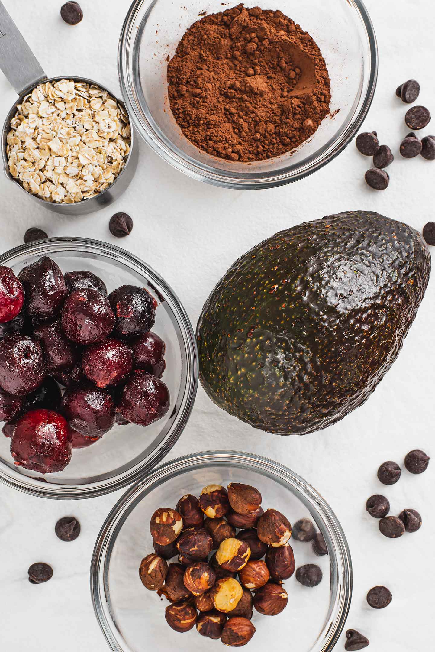 Top down view of an avocado, a dish of frozen cherries, a measuring cup of oats, and small dishes of cocoa powder and hazelnuts. Chocolate chips speckle the tray the ingredients sit upon.