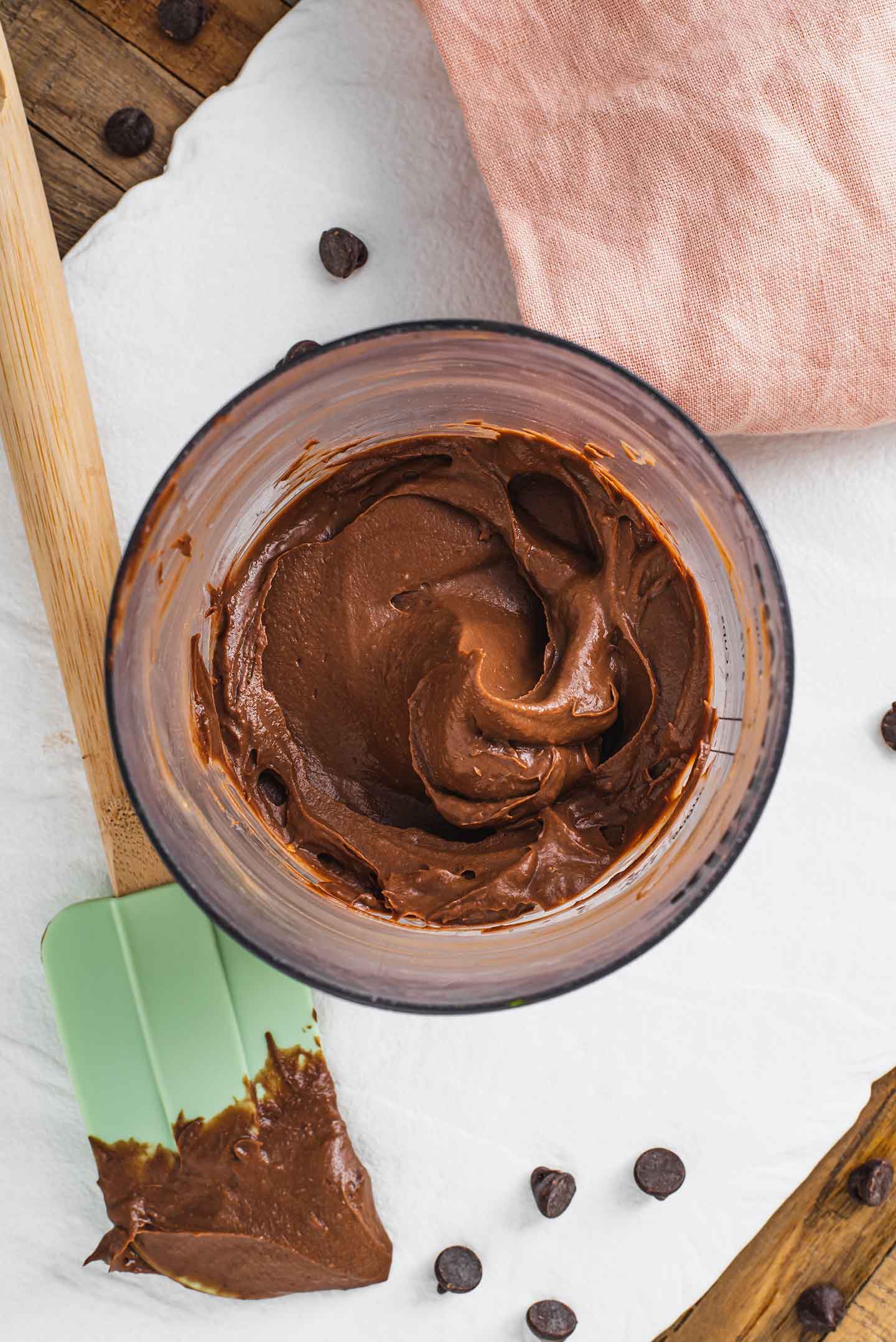 Top down view of thick chocolate avocado mousse in the cup of a hand blender. A rubber spatula with the thick mousse lays on a white tray.