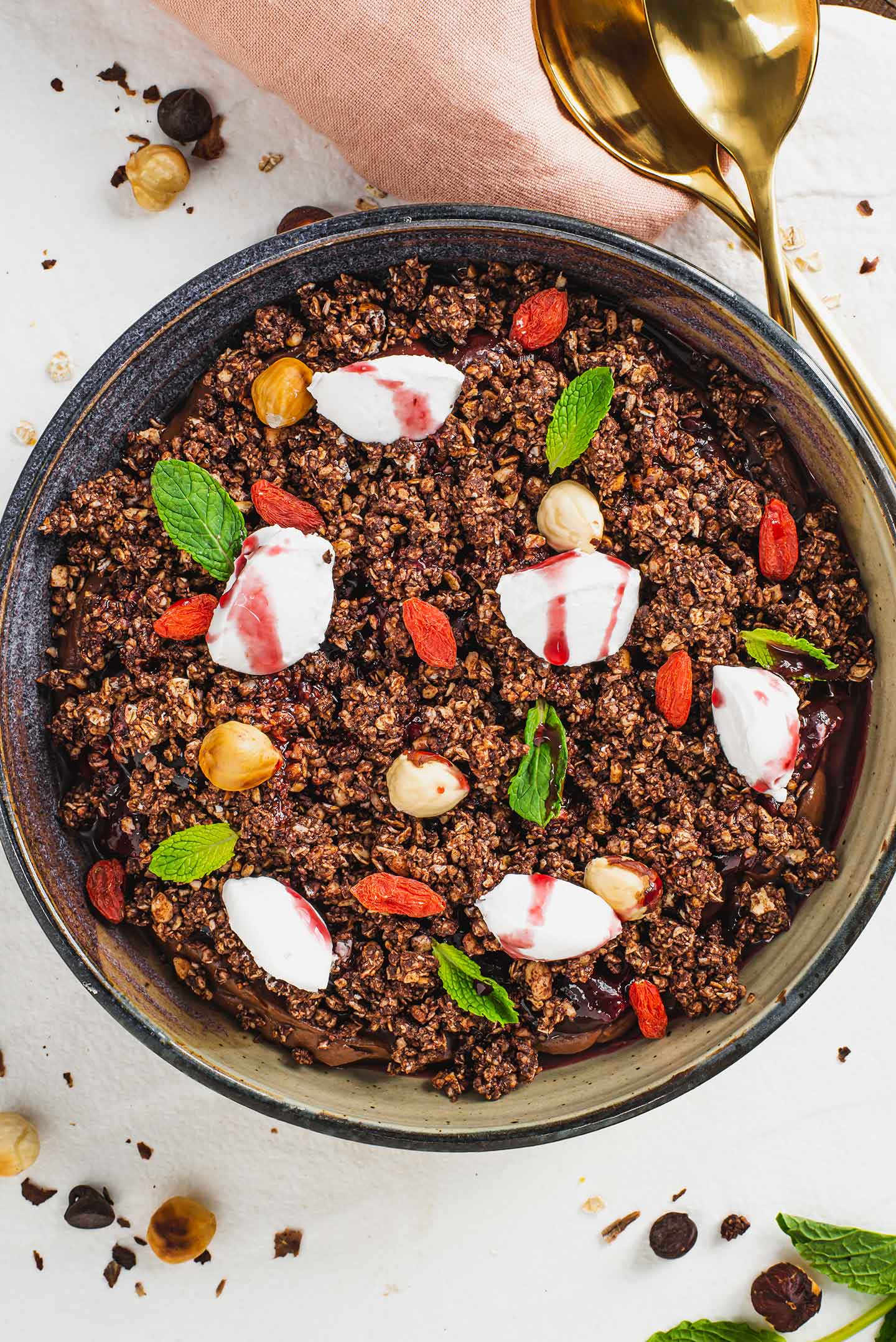 Top down view of a chocolate mousse terrarium. The top of the dessert is covered in an oat and nut crumble, dollops of whipped cream, dried goji berries, and mint leaves.