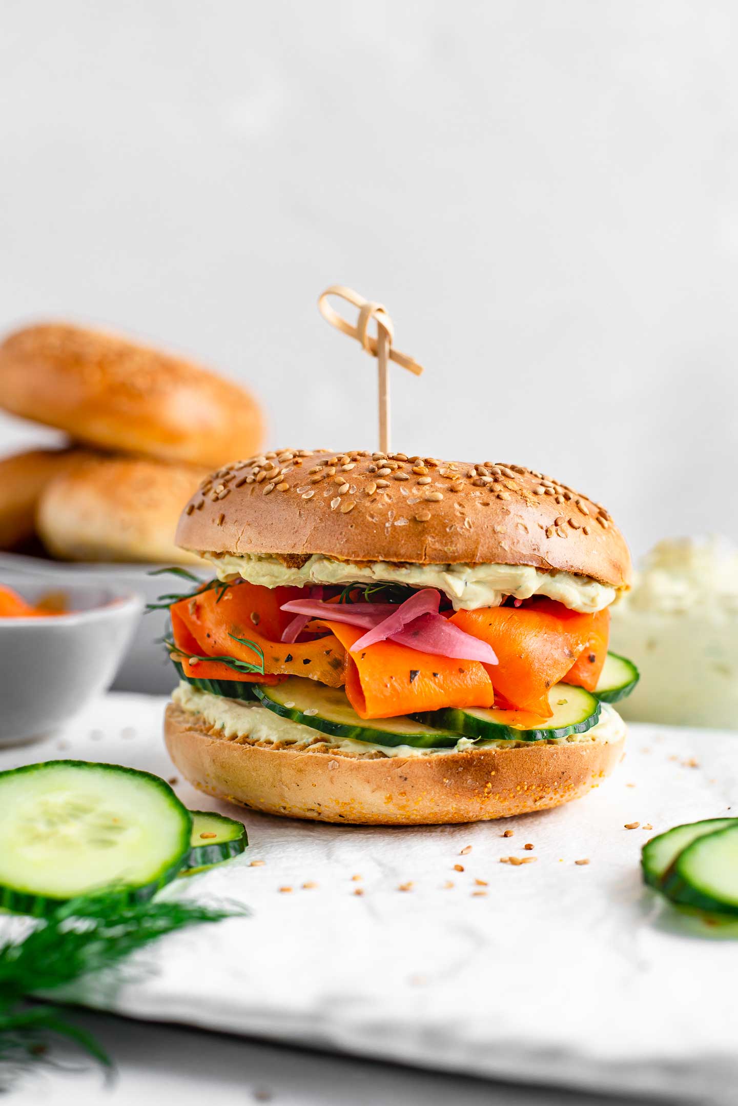 Side view of a loaded vegan carrot lox bagel sandwich. A toasted bagel stuffed with creamy dill spread, cucumber slices, smoky lox, dill and pickled red onion.