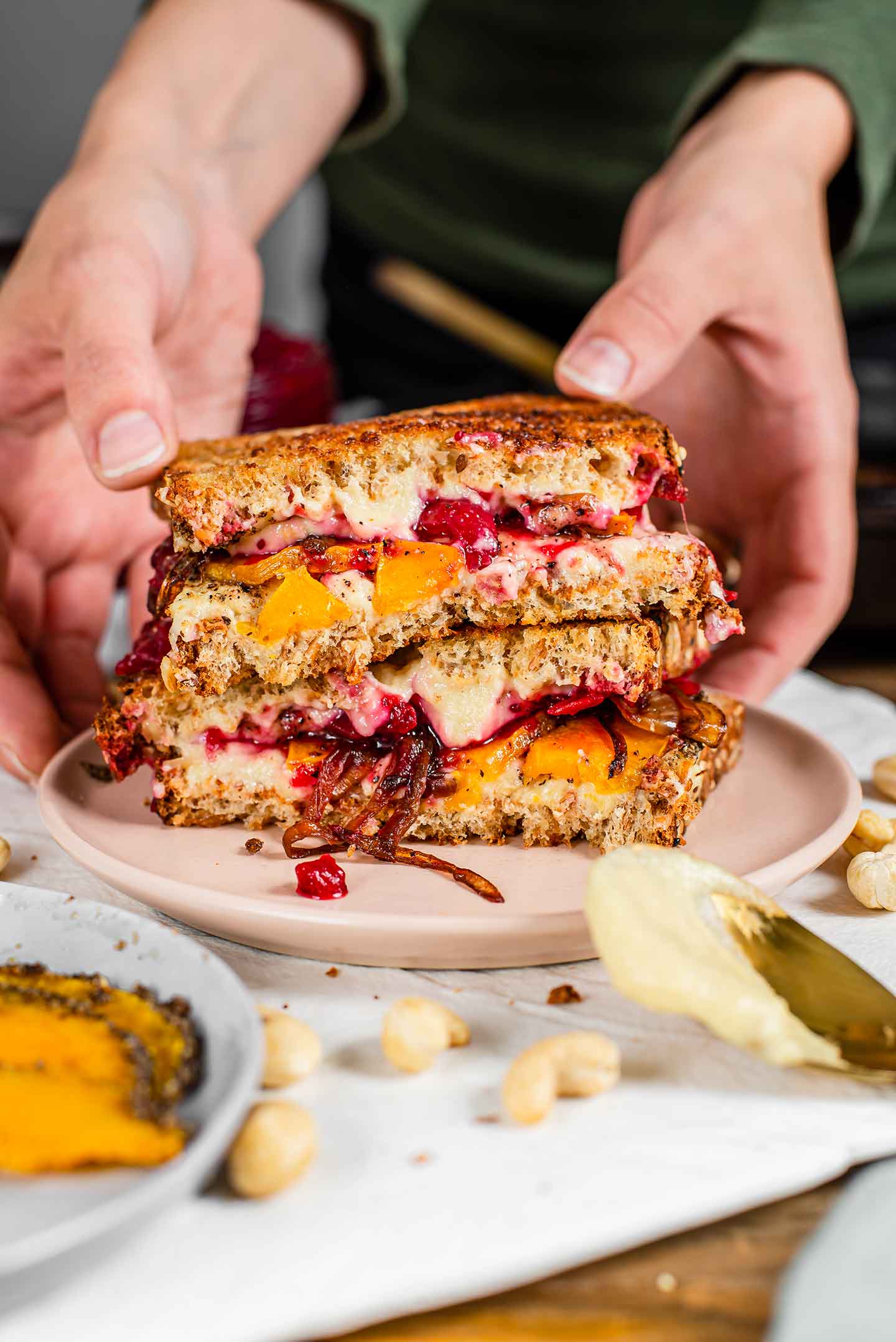 Side view of hands placing two halves of a sandwich on a plate. The sandwich is full of melty mozzarella, cranberry sauce, fried onions, and roasted squash.