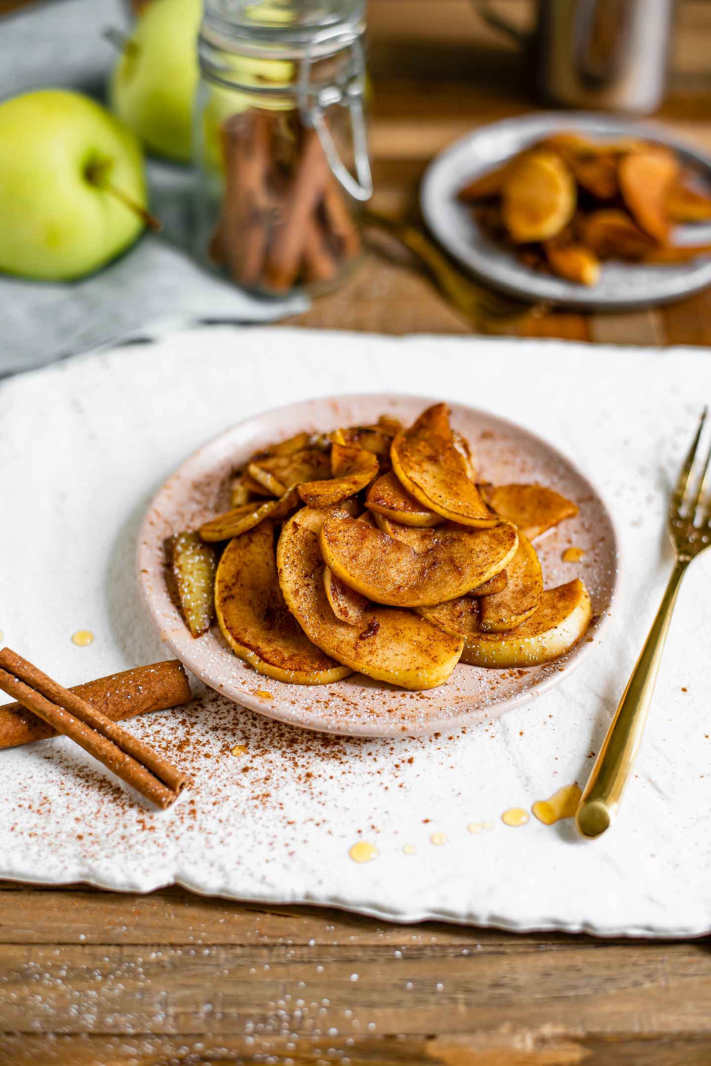 Warm, tender apple slices coated in cinnamon and a dusting of icing sugar are piled on a pink saucer. Apples, cinnamon sticks and another plate of warm cinnamon fried apples sit in the background. Cinnamon, maple syrup, and cinnamon is scattered around the plate.