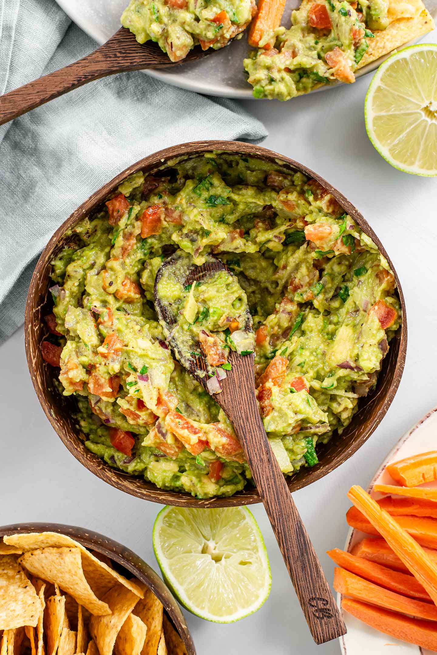 Top down view of a bamboo spoon in timely guacamole served in a coconut bowl. Tortilla chips, carrot sticks, and a squeezed lime surround the guacamole.