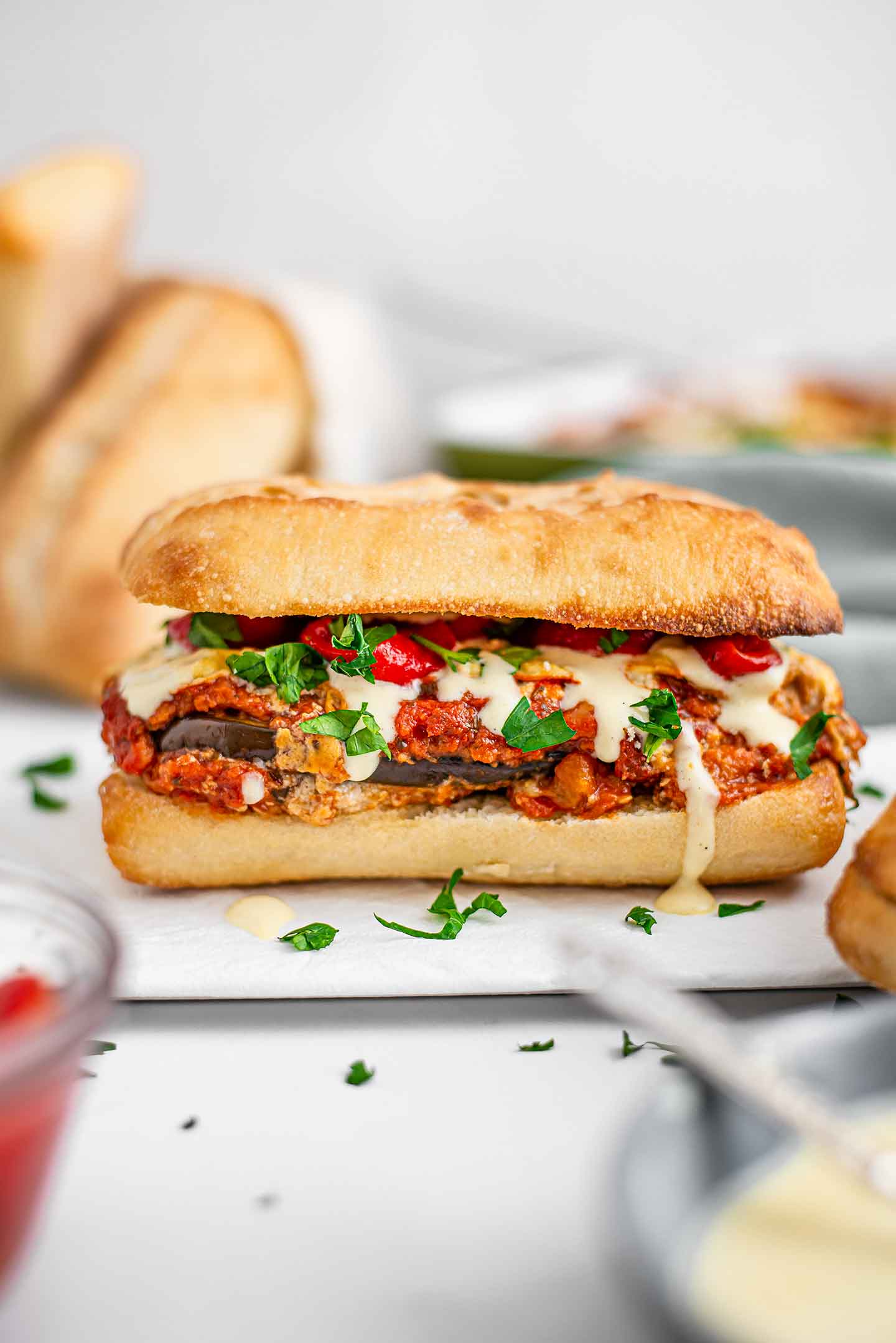A finished eggplant parmesan sandwich sits on a white tray with other buns, more eggplant, and more toppings around.