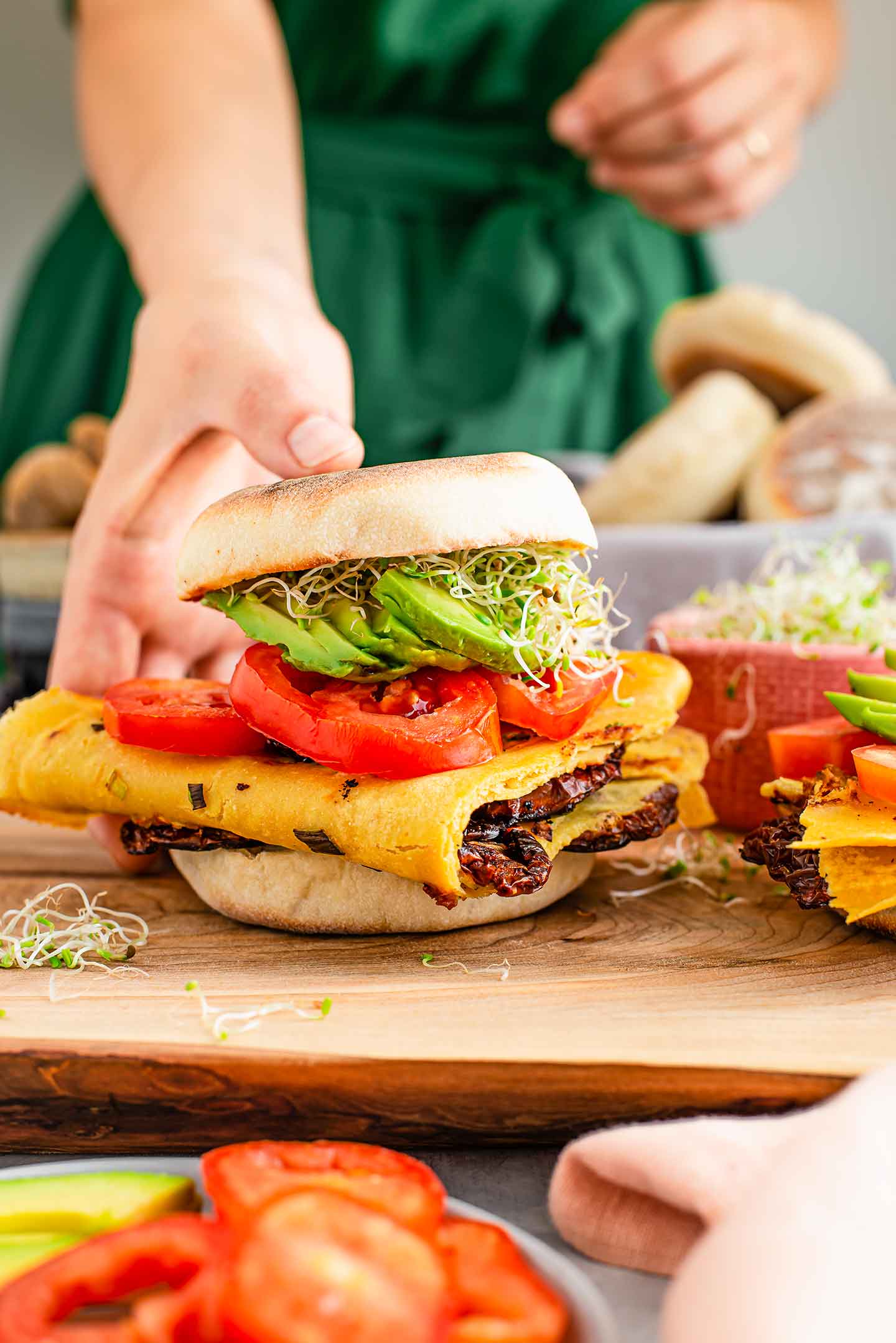 Side view of a towering vegan Portuguese breakfast sandwich. A chickpea omelette with vegan chourico is folded on a Portuguese muffin and topped with sliced tomato, avocado and alfalfa sprouts.