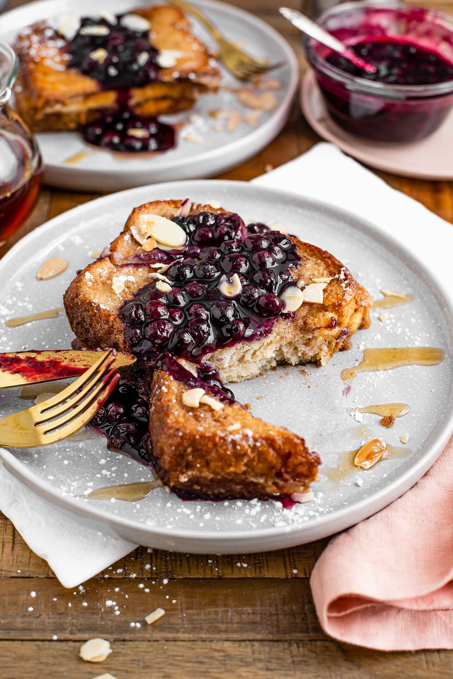 Side view of two golden slices of french toast stacked on one another, topped with a blueberry sauce, toasted sliced almonds, and drizzled with maple syrup.
