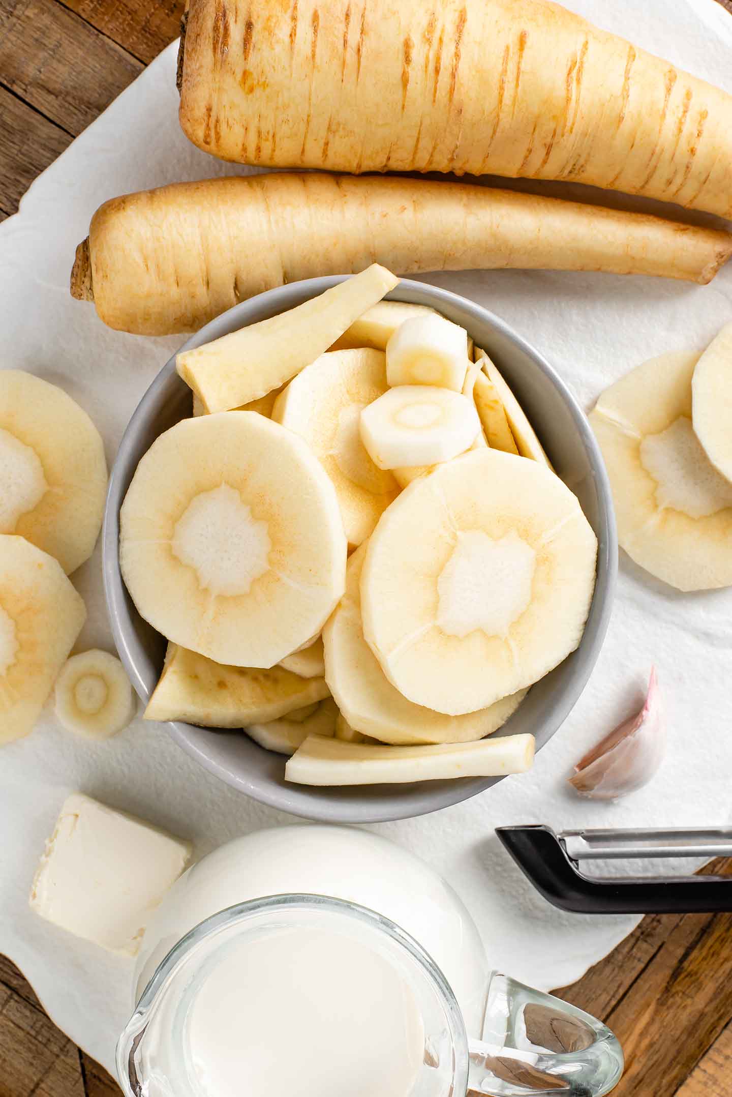 Top down view of ingredients for parsnip puree. Peeled and sliced parsnips are piled in a small bowl with garlic, butter, and milk surrounding.