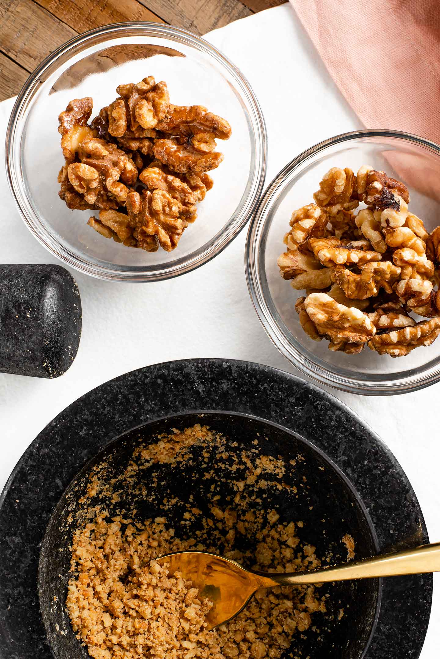 Top down view of crushed candied walnuts in a mortar and pestle. Raw walnuts and whole candied walnuts fill small dishes next to the mortar and pestle.