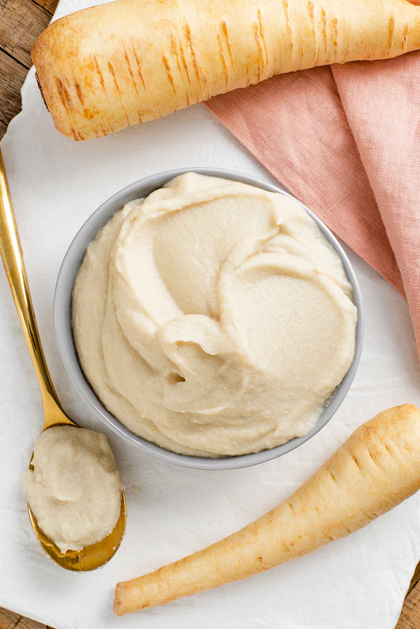 Top down view of a creamy parsnip puree filling a small bowl. A spoon with the creamy mixture lays beside the bowl and whole parsnips are on the edge of the photo.