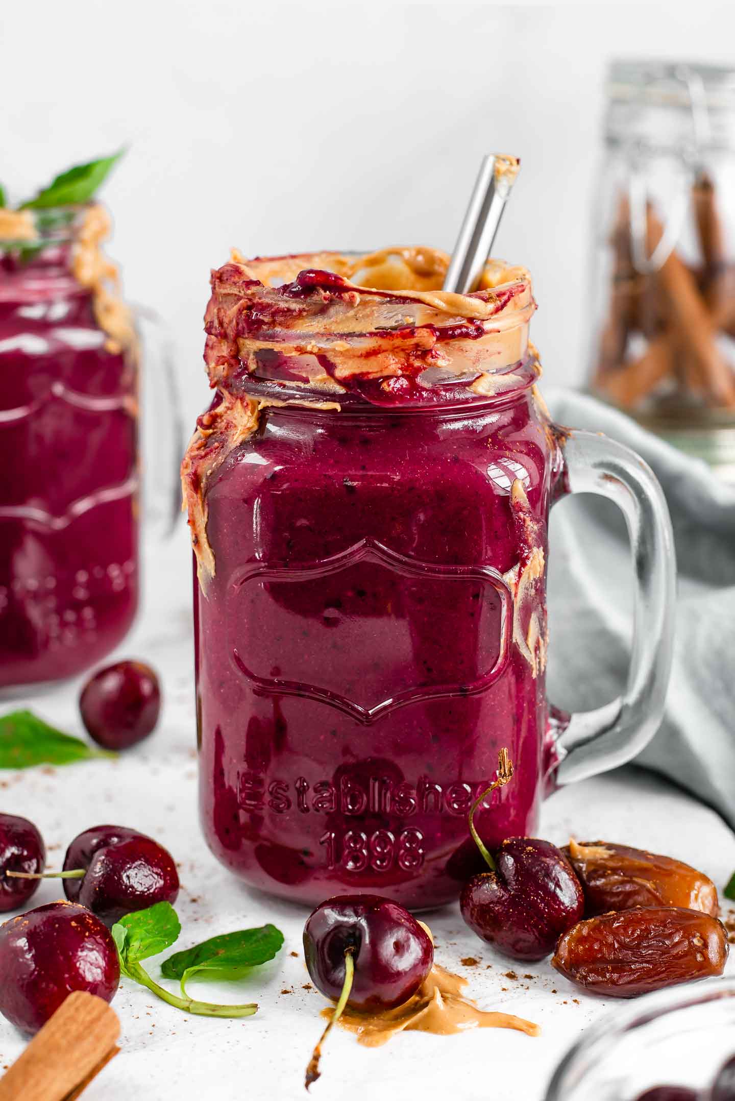 Side view of chocolate beet smoothie in a glass jar smeared with peanut butter. Another smoothie fills the background and cherries, dates, cinnamon sticks, and mint leaves are scattered around.