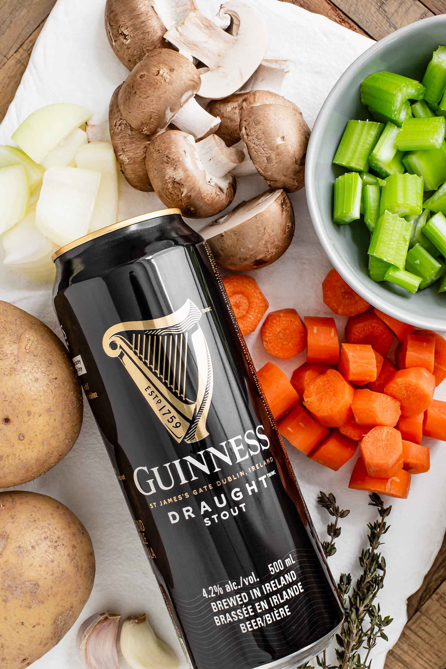Top down view of ingredients. A can of Guinness stout is surrounded by carrots, celery, mushrooms, onion, potatoes, garlic and fresh thyme.