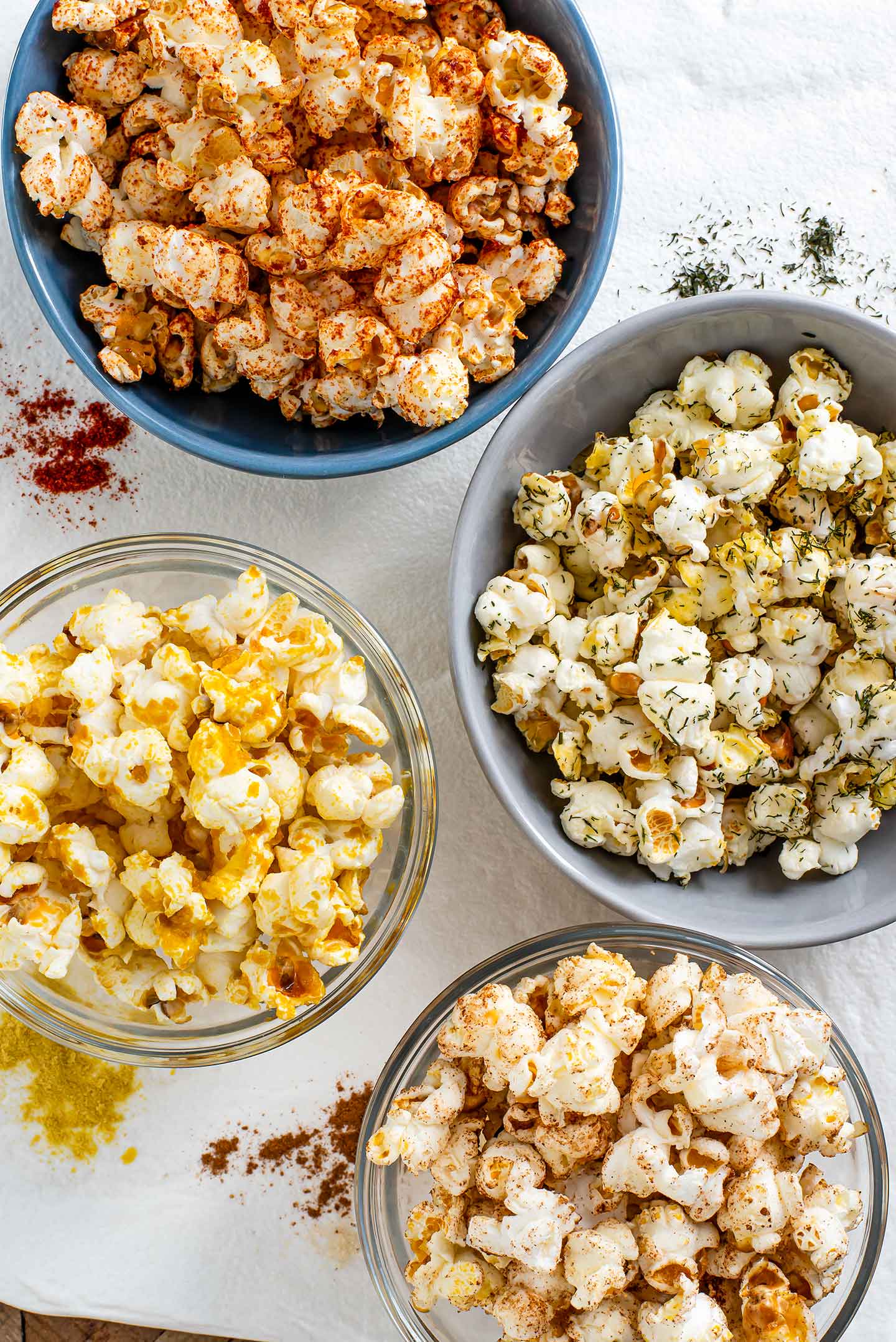 Top down view of four small bowls with different flavoured popcorn. The corresponding seasonings are spread on a white tray next to the bowls.