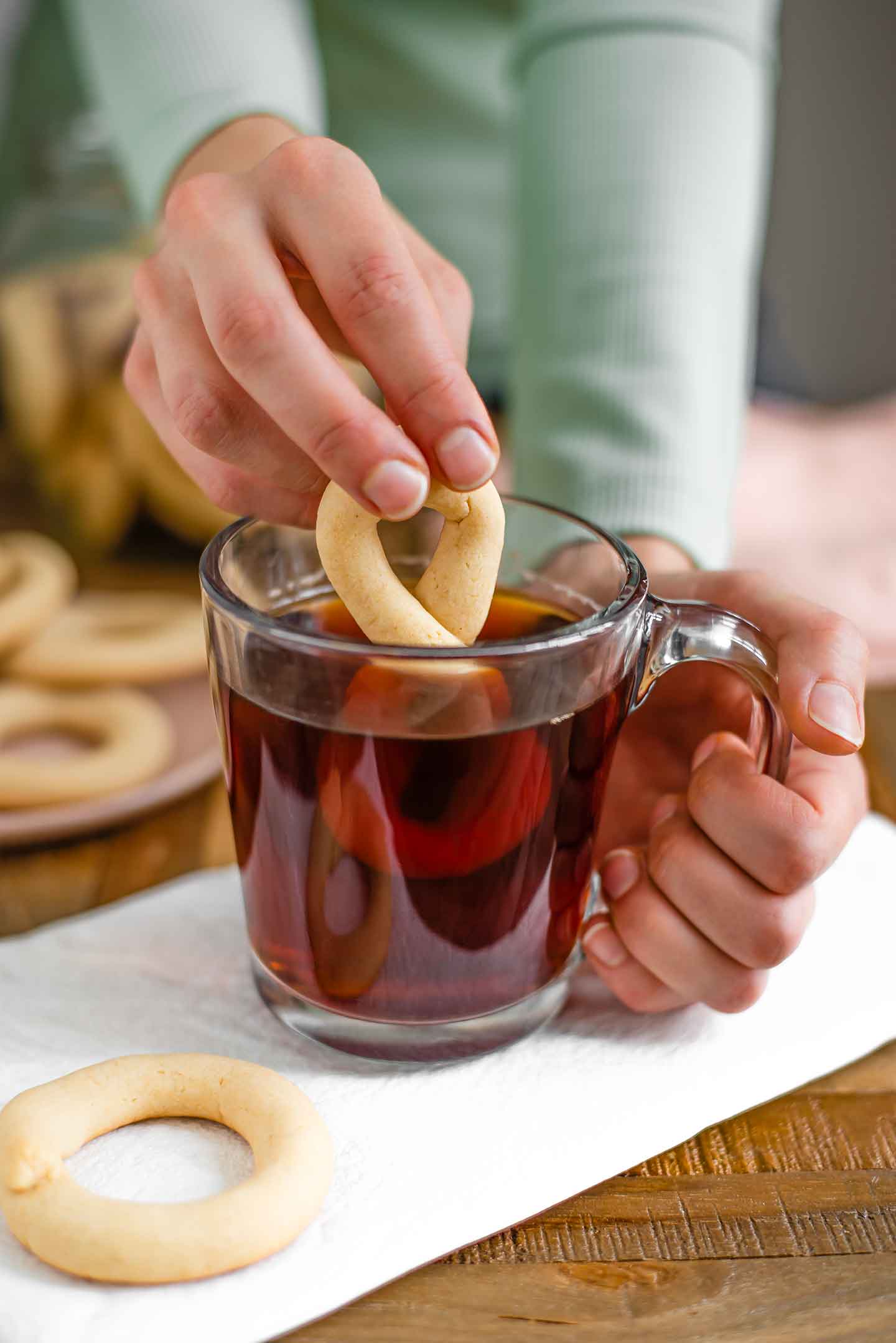 Side view of a hand dunking a golden figure eight Portuguese cookie into a mug of tea. Other cookies are scattered around.