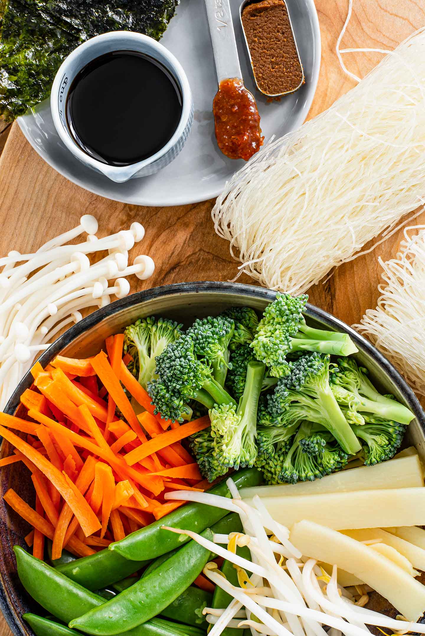 Top down view of ingredients. Tamari, miso paste, and chili garlic sauce are pictured amoung dry rice noodles, enoki mushrooms, carrots, broccoli, snap peas, bamboo shoots, and bean sprouts.