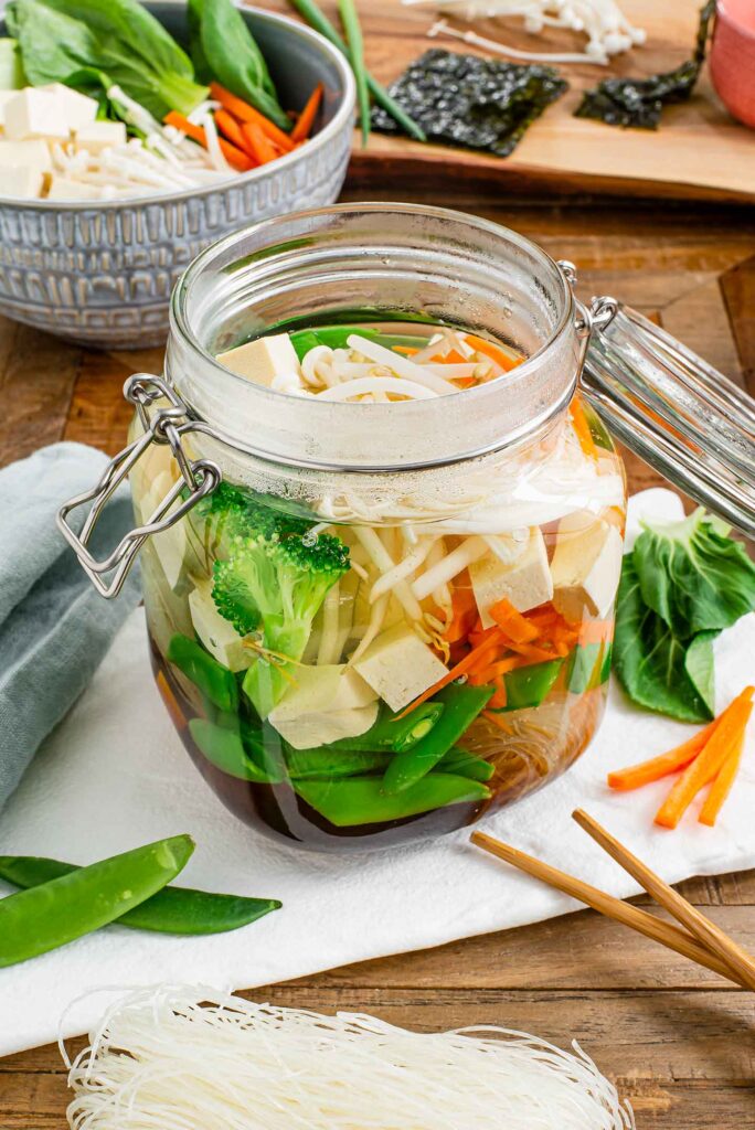 Side view of a glass jar with a clamp lid filled with vegetables, tofu, rice vermicelli noodles, and a dark broth. Chop sticks and extra ingredients surround the jar of soup.