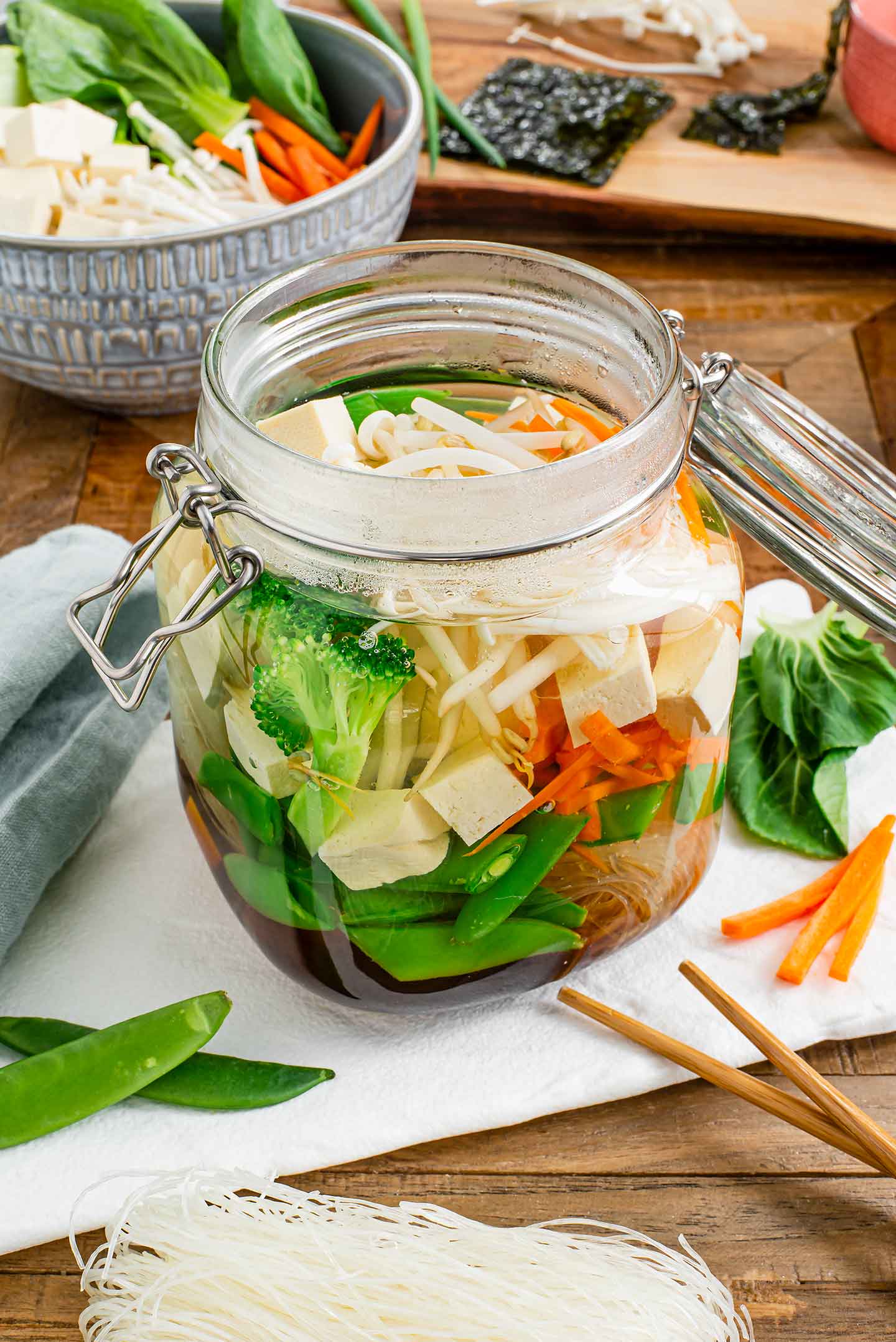 Side view of a glass jar with a clamp lid filled with vegetables, tofu, rice vermicelli noodles, and a dark broth. Chop sticks and extra ingredients surround the jar of soup.