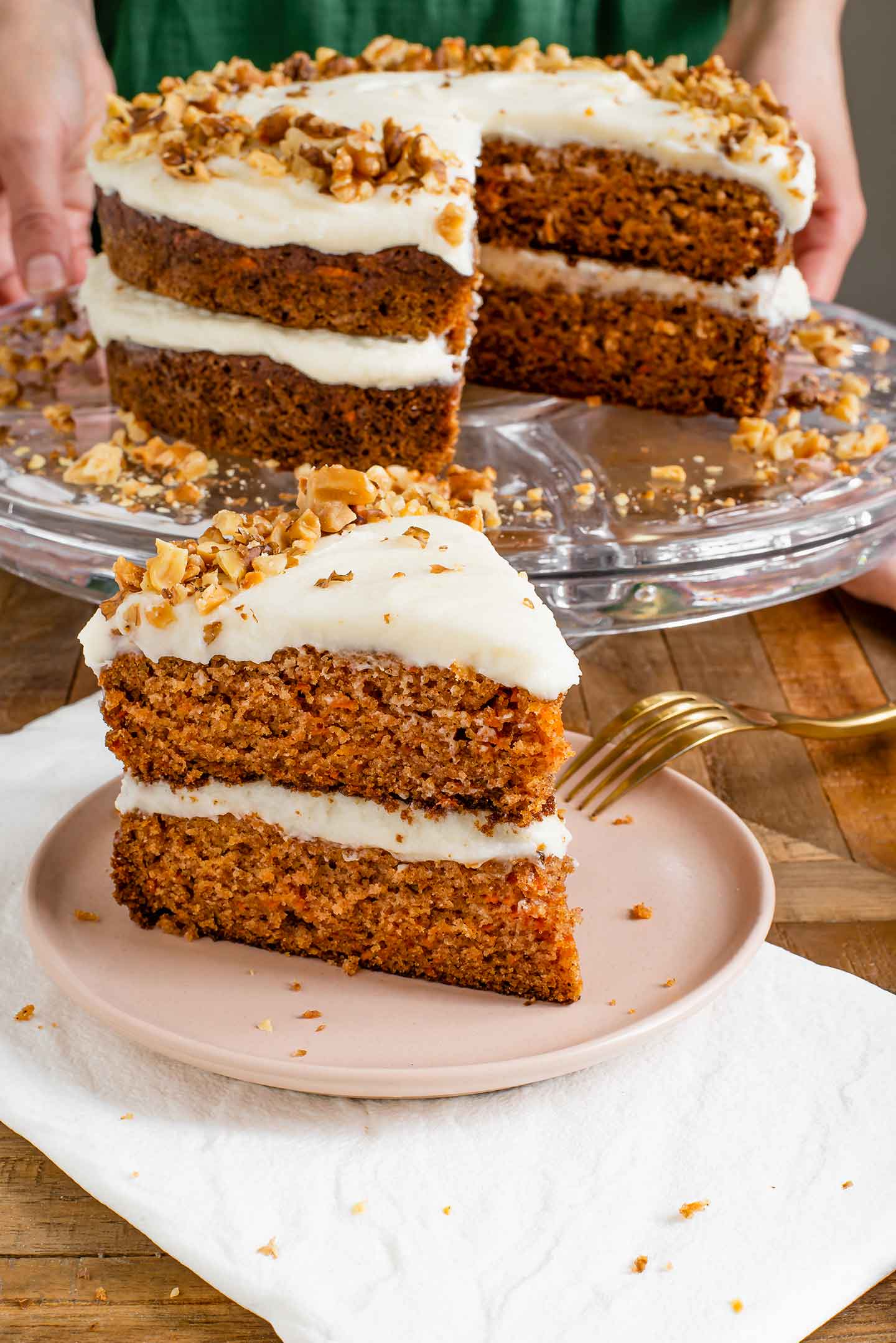 Side view of a large slice of vegan carrot cake with dairy-free cream cheese frosting. The rest of the cake is on a cake stand in the background.