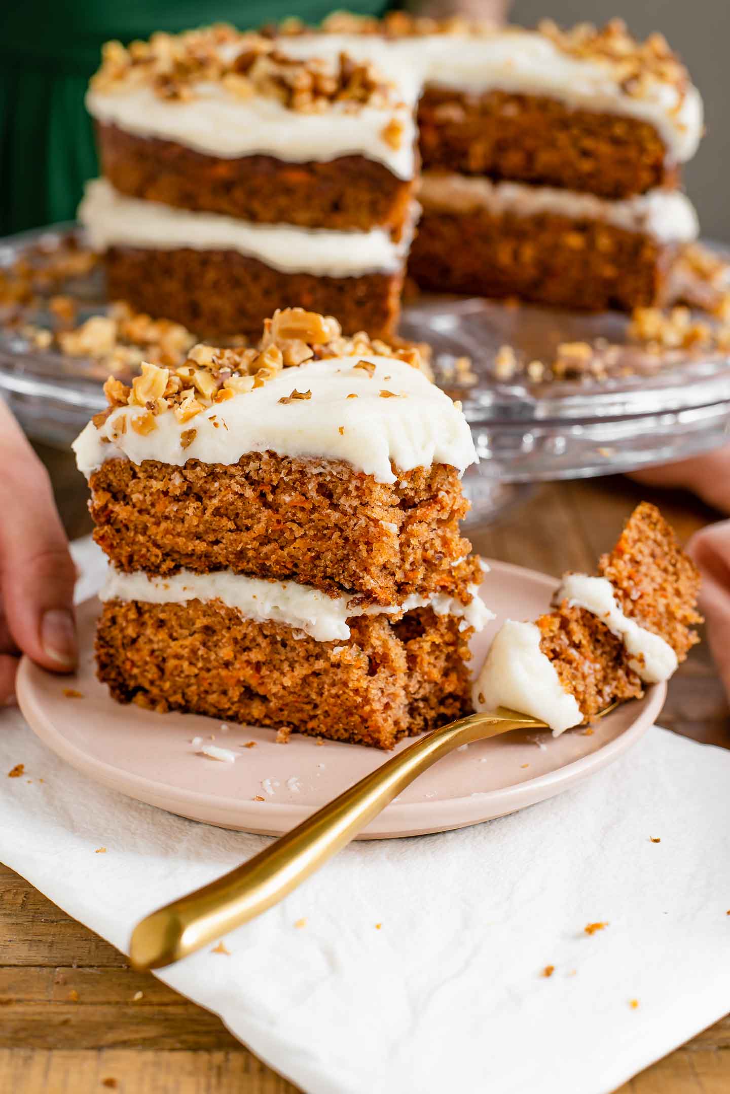 Side view of a large slice of a two tiered carrot cake with cream cheese frosting. A fork has sliced through the piece and lays on the plate.