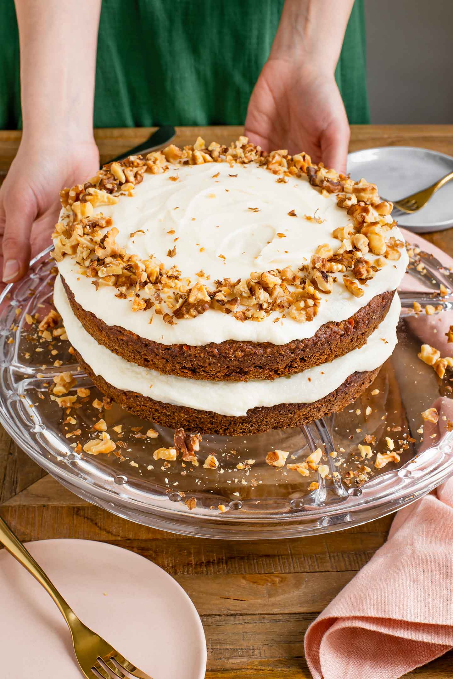 A two tier vegan carrot cake is presented on a cake stand. The sides of the cake are not iced but the centre and top is frosted with vegan cream cheese frosting and decorated with crushed toasted walnuts.