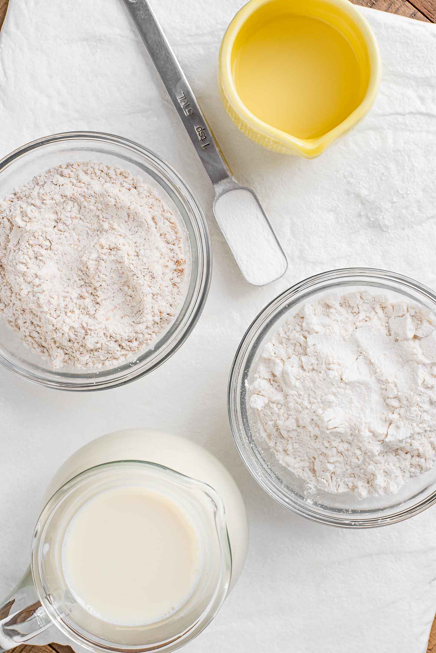 Top down view of ingredients on a white tray. Small bowls of white and whole wheat flour, soy milk, lemon juice, salt, and a teaspoon of baking soda are pictured.