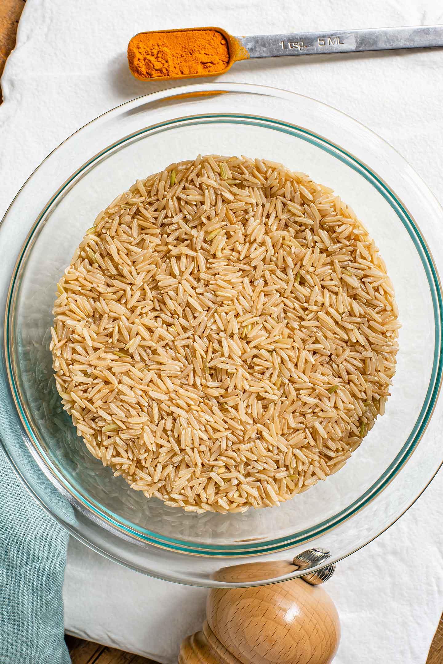 Top down view of uncooked brown jasmine rice in a bowl with a teaspoon of turmeric powder and a salt mill nearby