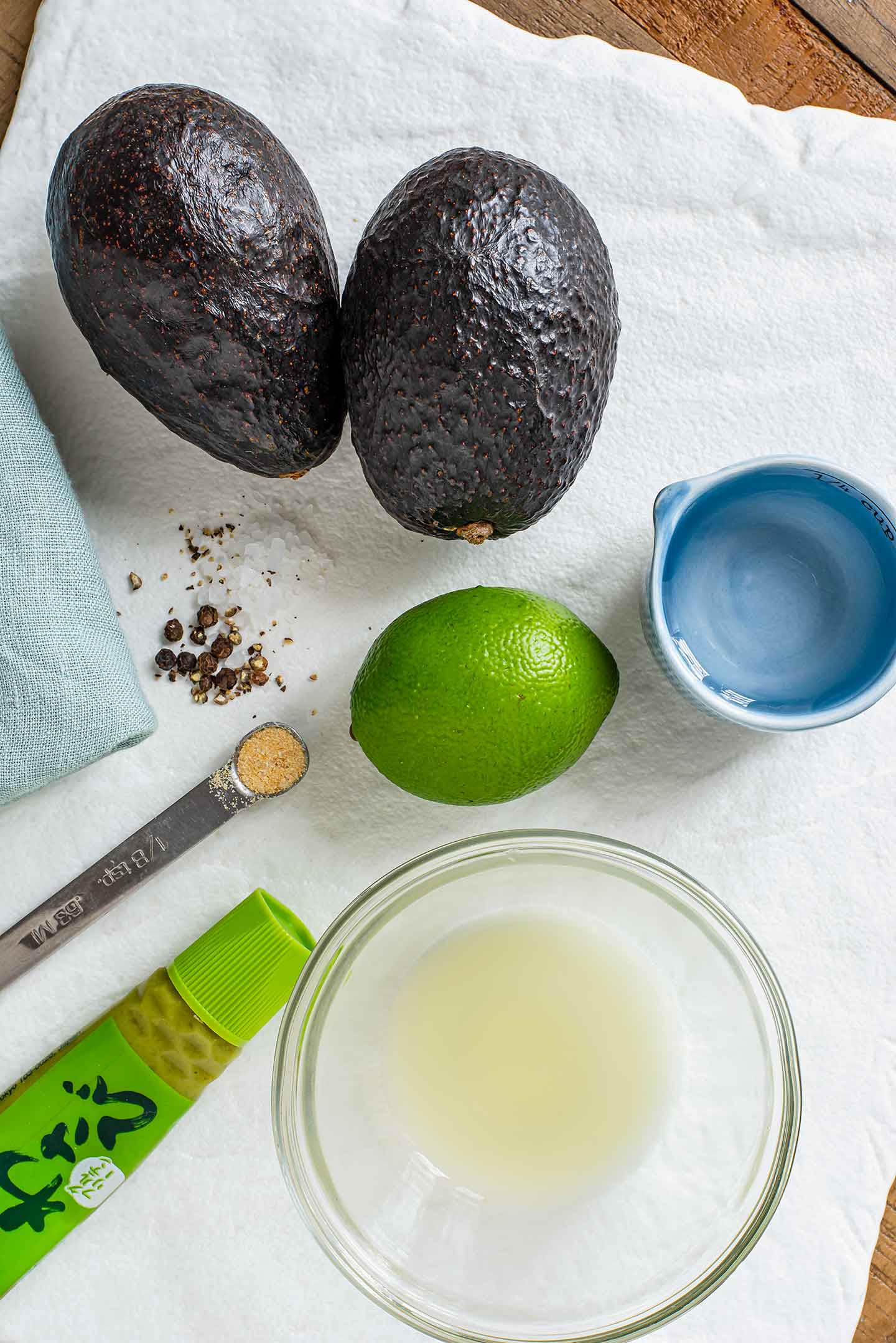 Top down view of ingredients on a white tray. Two small avocados, a lime, lime juice, peppercorns, garlic powder, water, and wasabi.