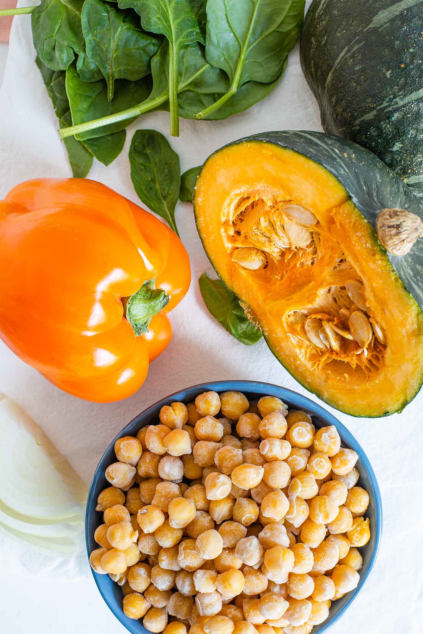 Top down view of a small kabocha squash sliced in half and surrounded by cooked chickpeas, an orange bell pepper, and baby spinach leaves.