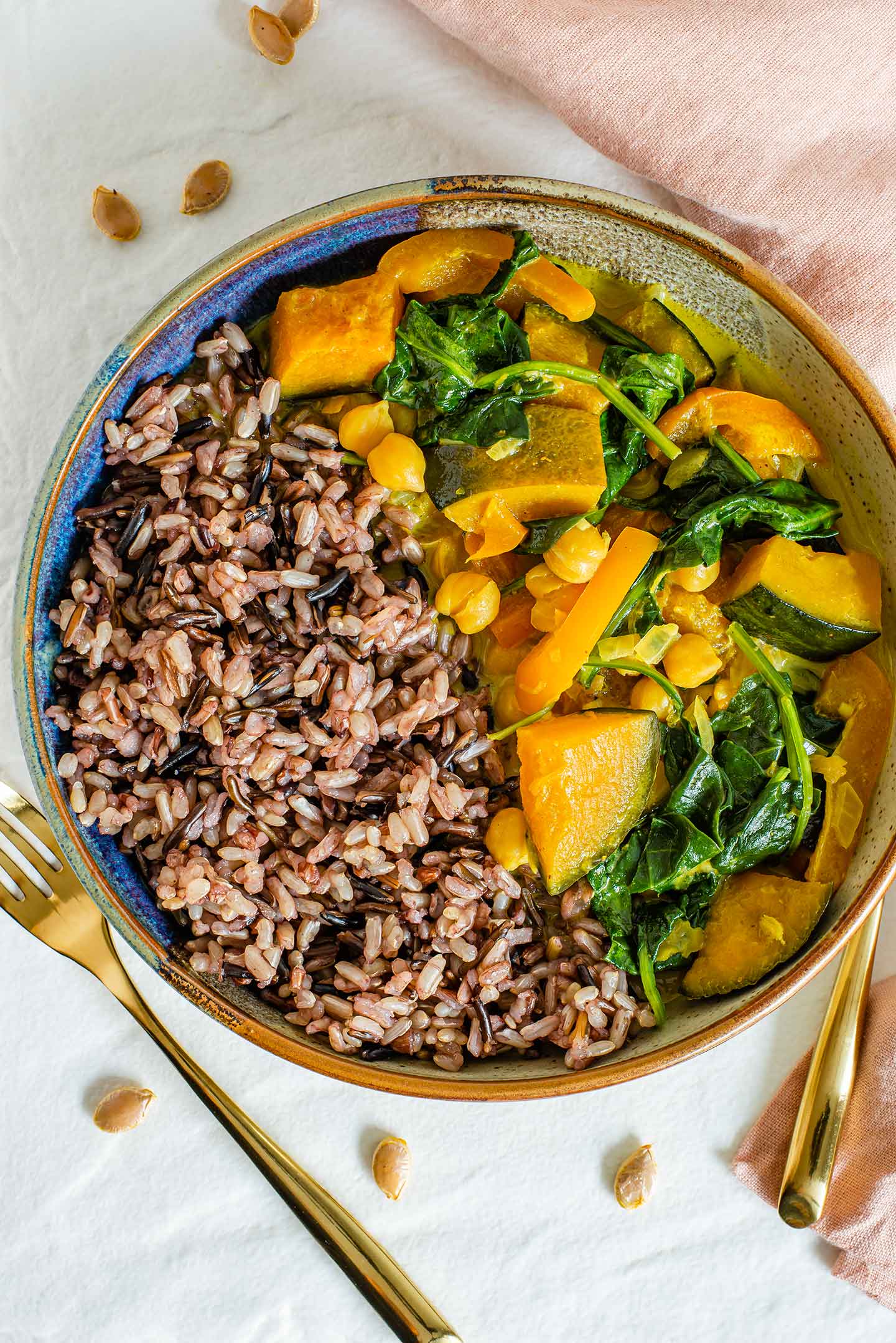 Top down view of a creamy curry with chunks of kabocha squash, chickpeas, and spinach in a bowl with a side of wild rice.
