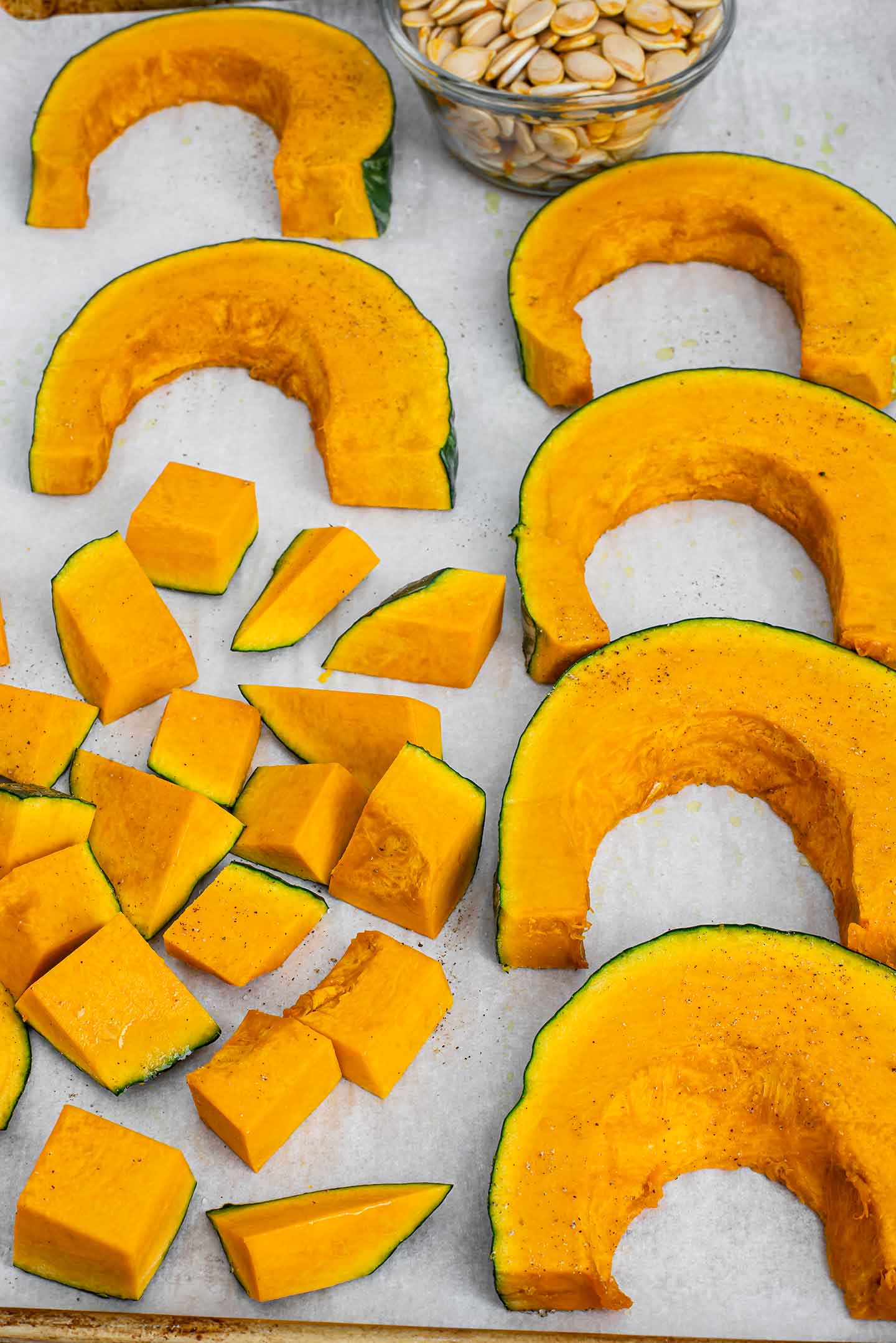 Sliced and cubed squash lays on a baking tray lined with parchment paper. Cleaned seeds fill a small bowl and the peel remains on the squash.