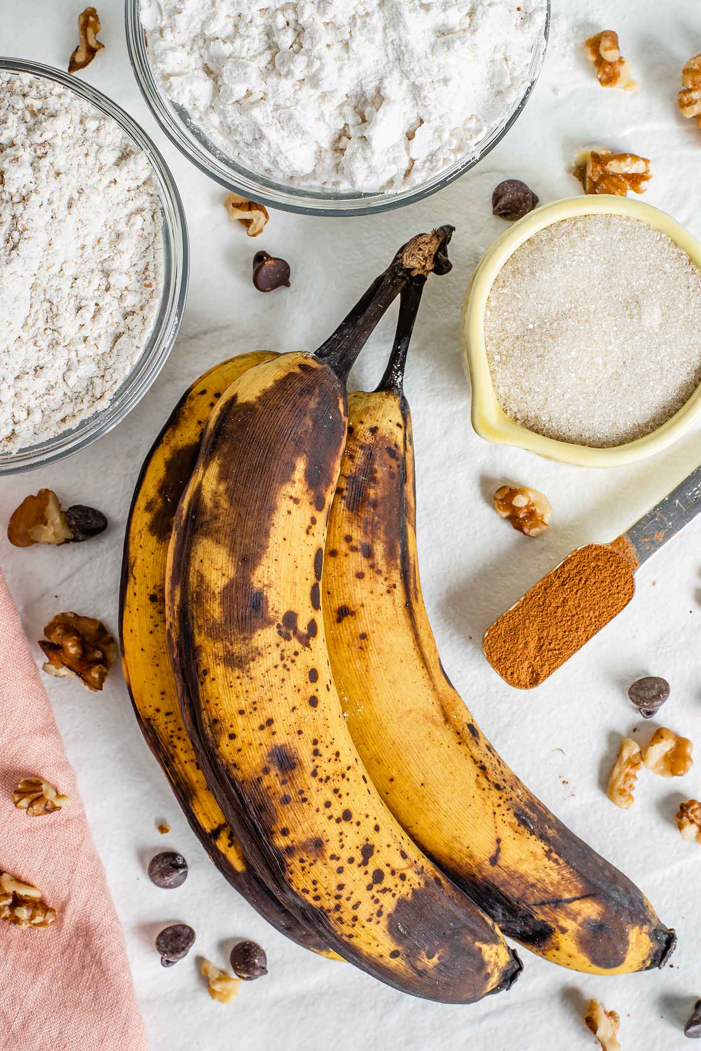 Top down view of ingredients on a white tray. Three overripe bananas, white and whole wheat flour, cane sugar, and cinnamon are pictured.